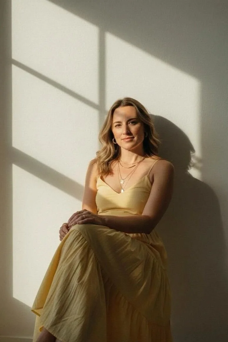 A woman with light brown hair wearing a sleeveless brown outfit with layered necklaces, posing with arms crossed against a dark background with sunlight casting shadows.