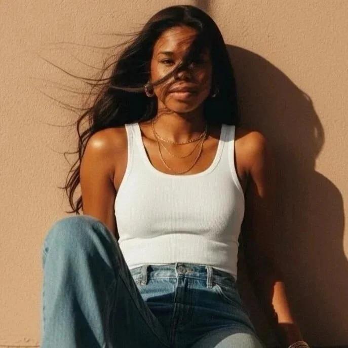 A young woman sitting against a beige wall, with wind blowing her long hair, dressed in a white tank top and blue jeans, bathed in sunlight.