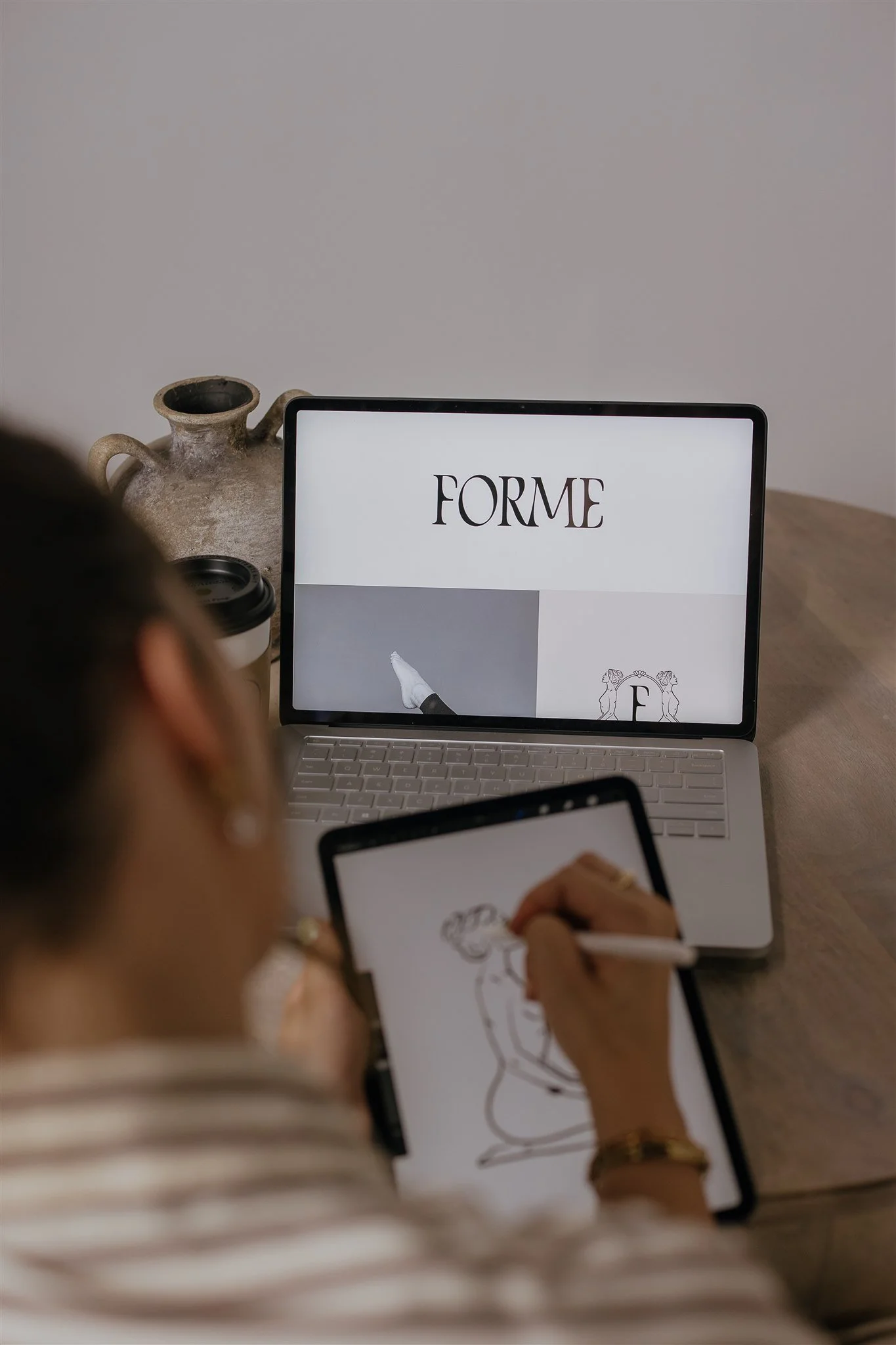 A woman drawing a fashion figure on a digital tablet with a stylus, with a laptop open in front of her displaying the word 'FORME' and a logo.