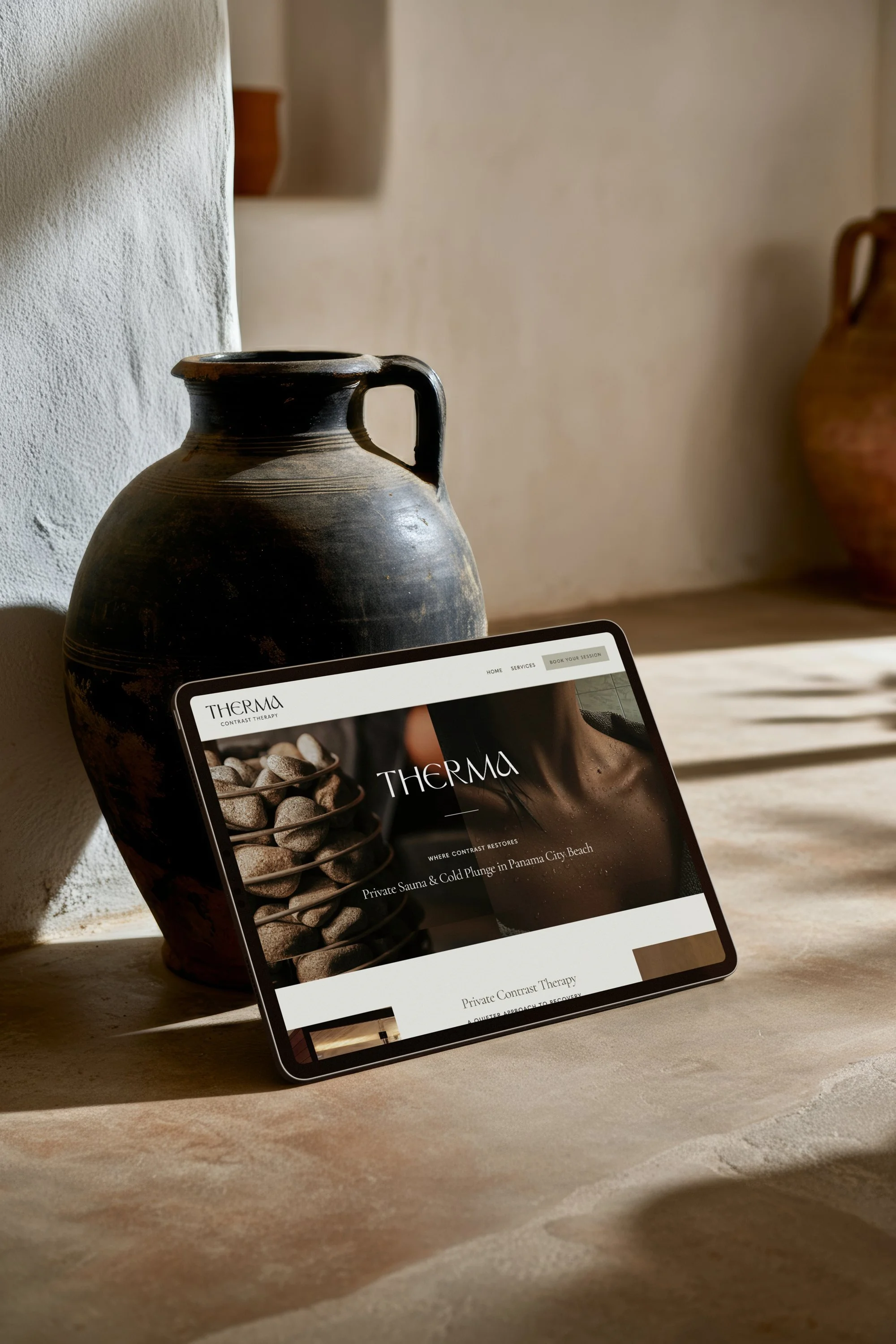A tablet displaying the website for a wellness retreat called Therma, showing a close-up of a woman's neck and a container of stones, placed on a wooden surface next to a large, antique ceramic vase.