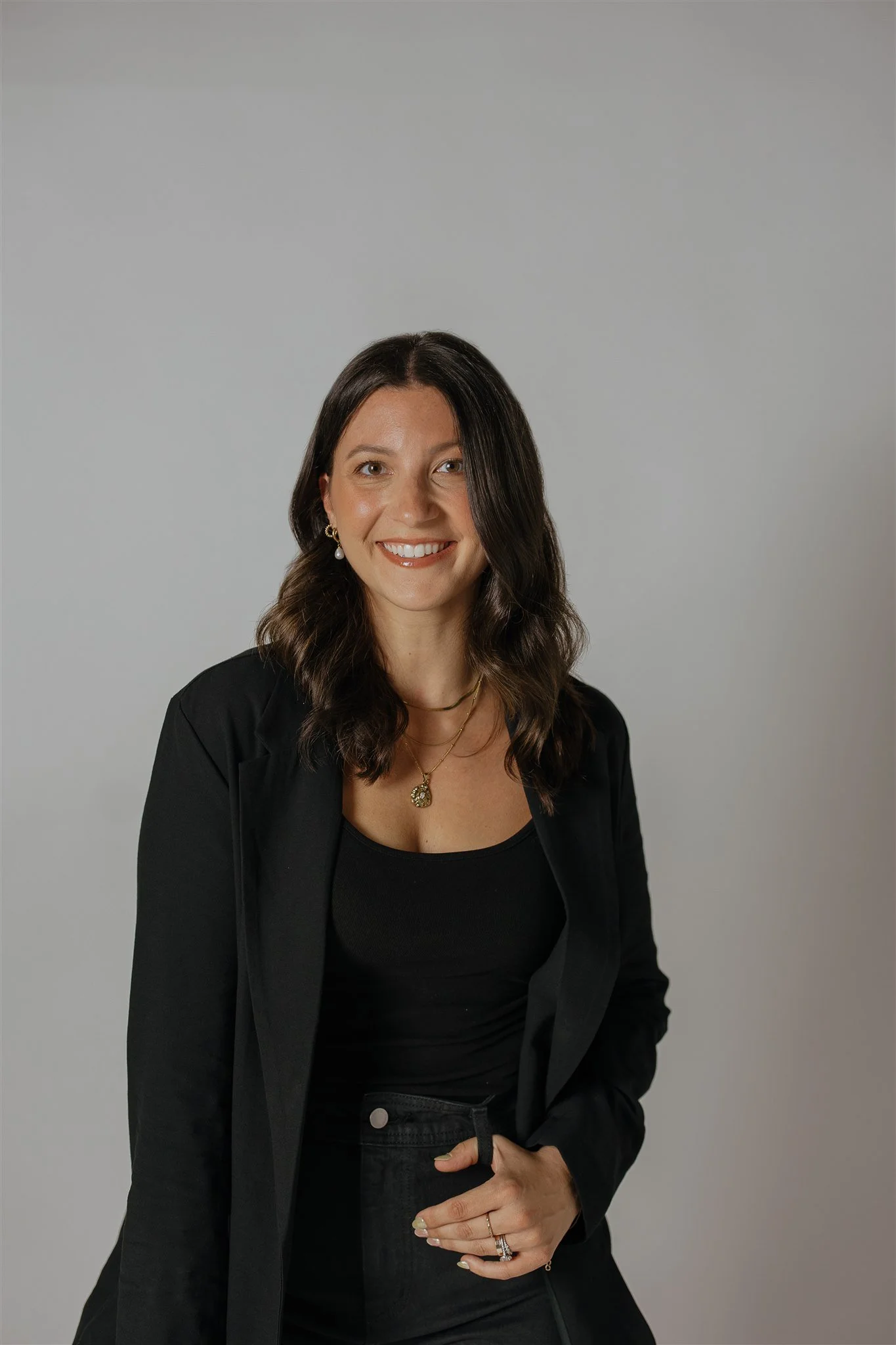 A woman with dark wavy hair wearing a black blazer and jewelry, smiling against a plain light gray background.