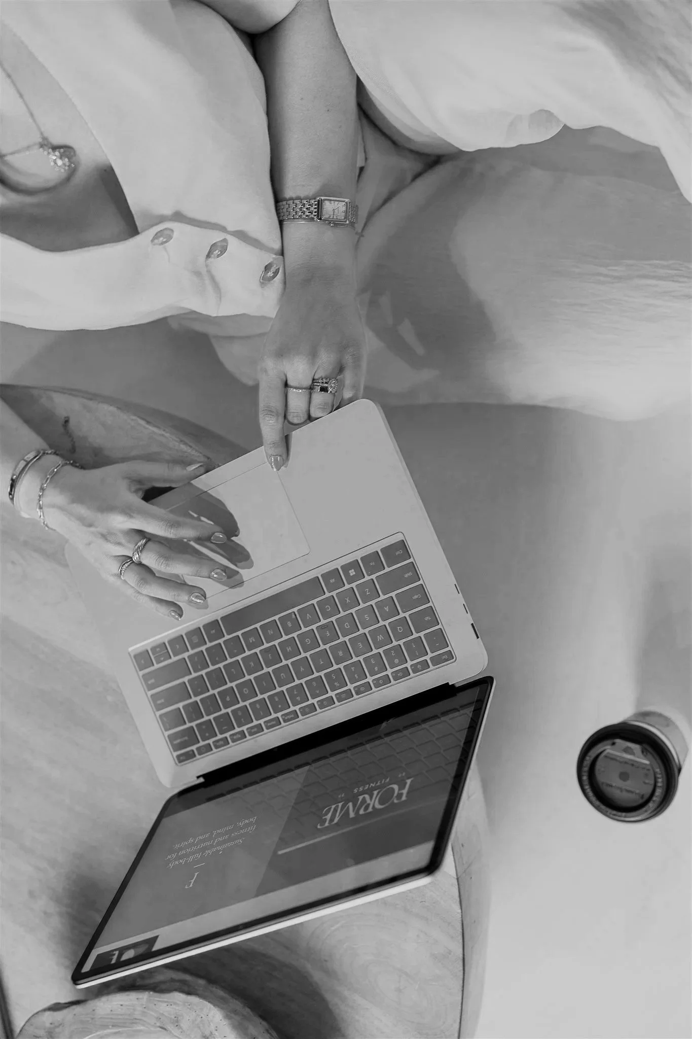 Top-down view of a luxury brand & web designer working on a laptop with a coffee drink nearby, wearing rings and bracelets, and a light-colored shirt.