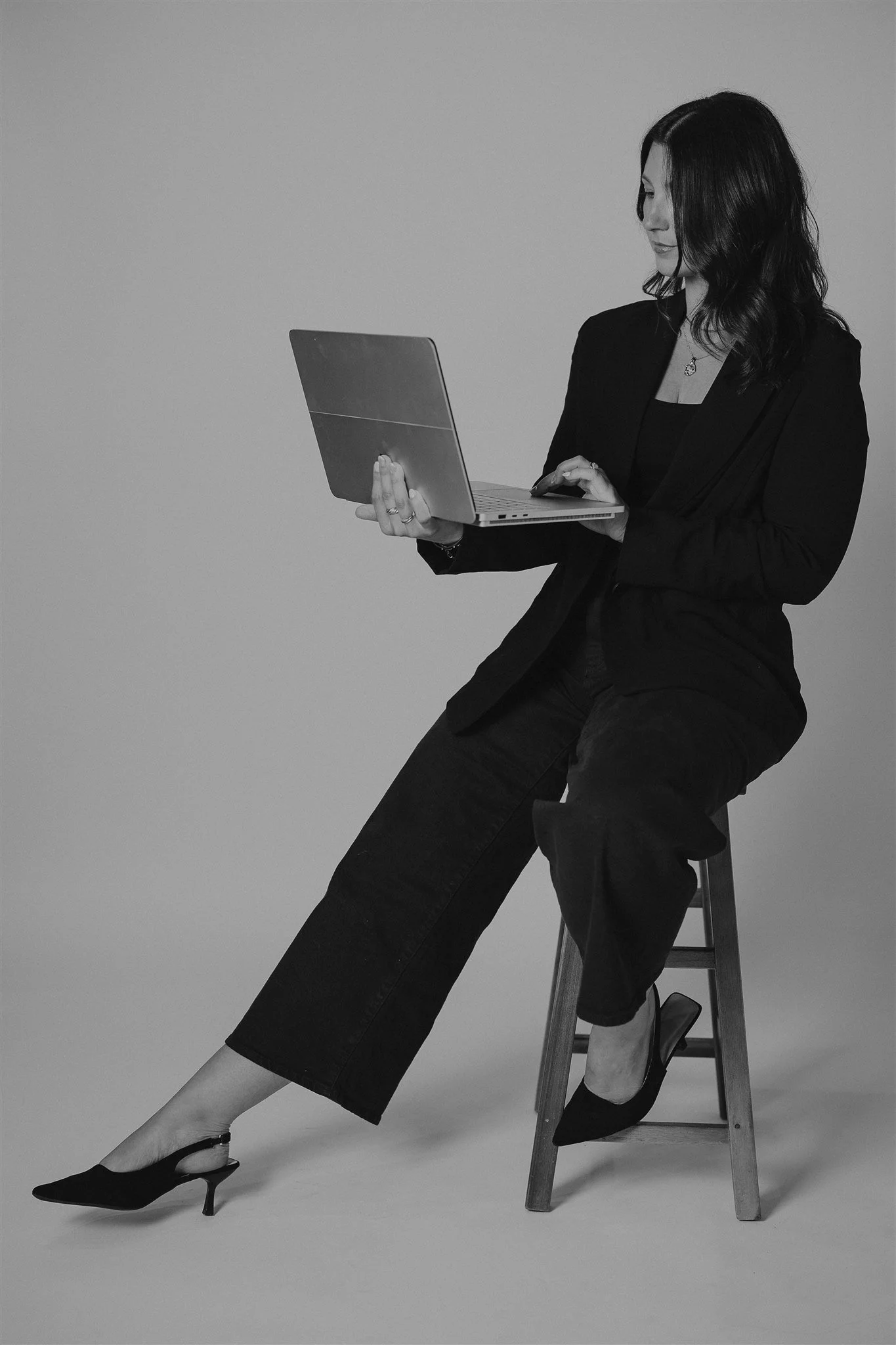 A woman in formal business attire, sitting on a wooden stool, holding a laptop in her left hand and using a mouse with her right hand.