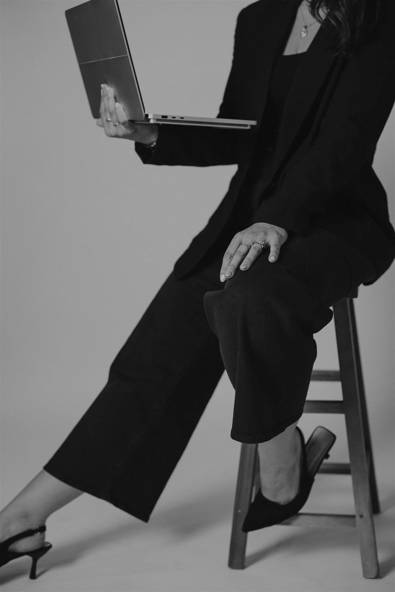 A woman dressed in black sitting on a stool, holding an open laptop.