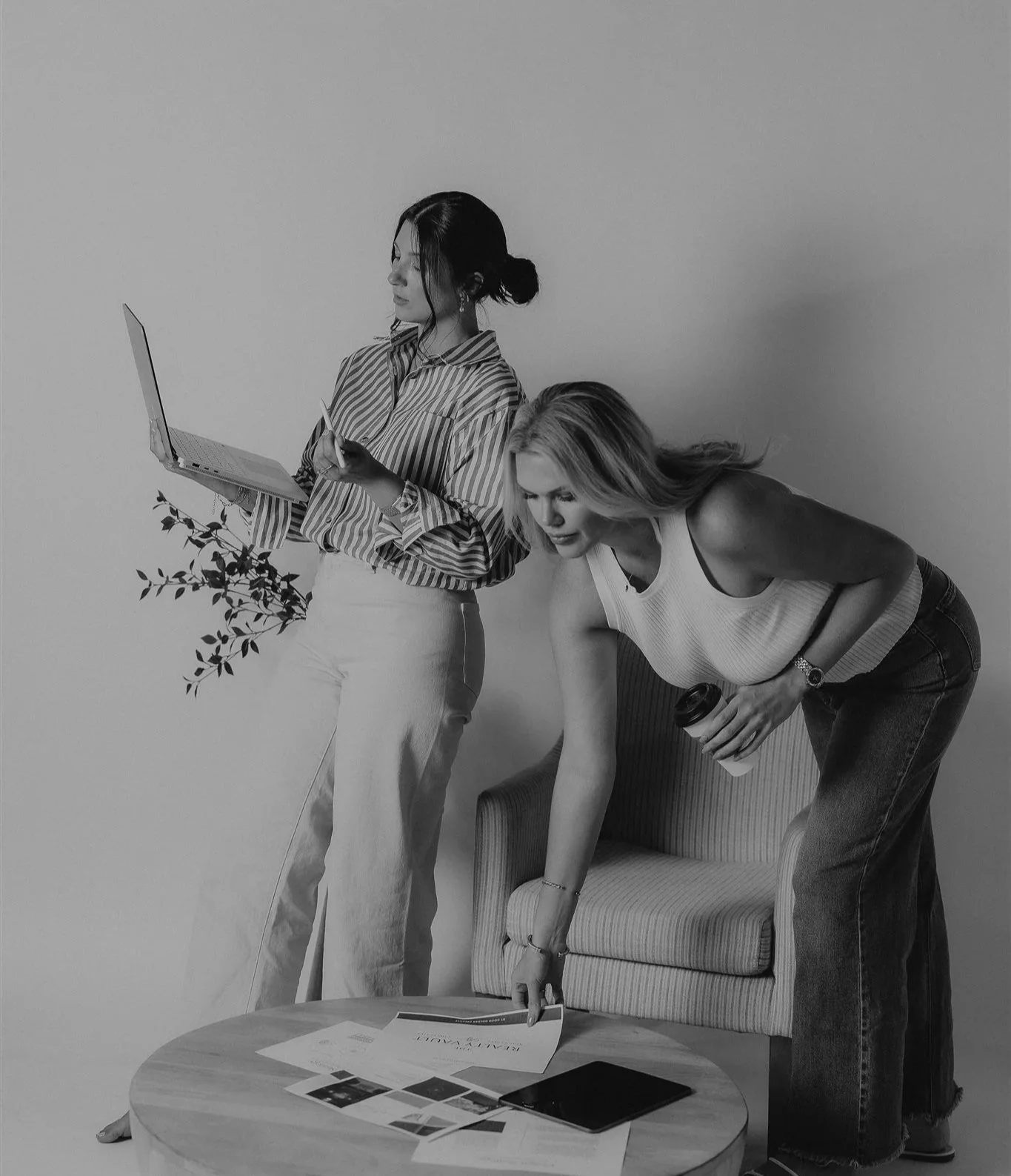 Two women working together in an office setting; one is standing with a laptop and notes, the other is leaning over a table with papers and a coffee cup.