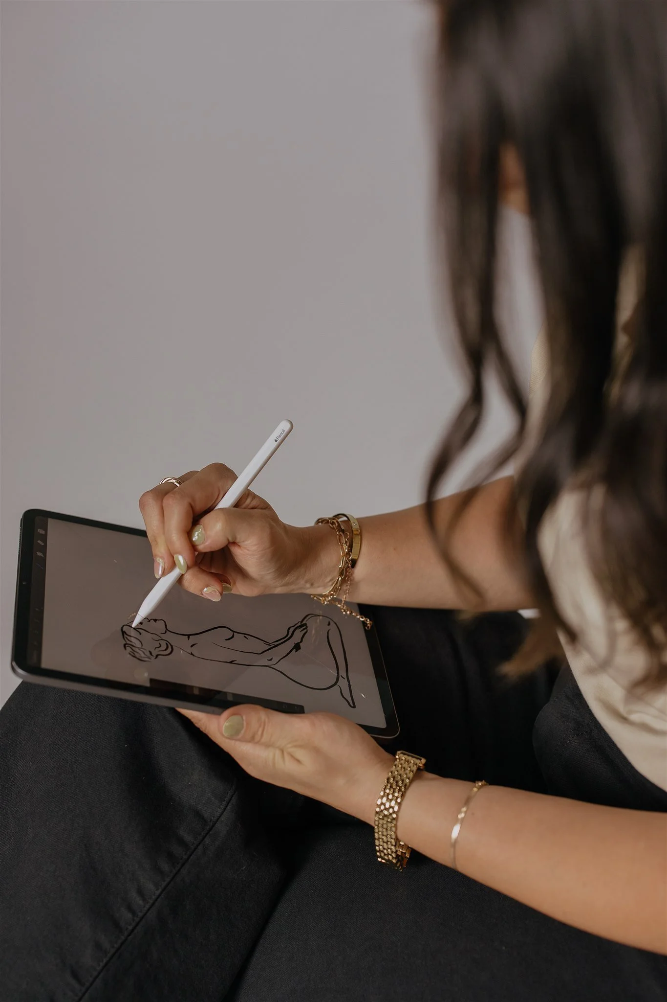 A woman with curly hair using a stylus pen to draw a line art illustration of a seated woman on a digital tablet. She is wearing jewelry including bracelets and rings.