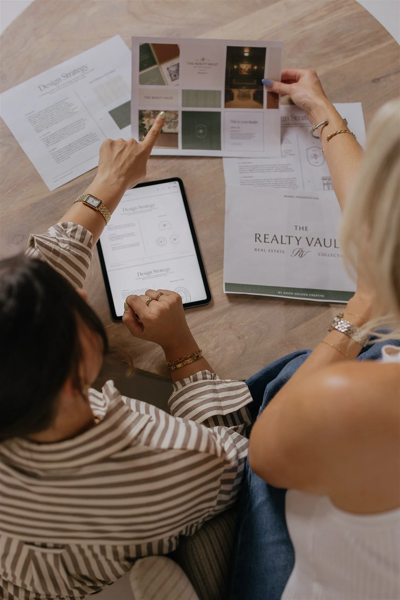 Two women reviewing printed and digital real estate marketing materials on a wooden table, with a tablet displaying a design strategy document and some brochures.