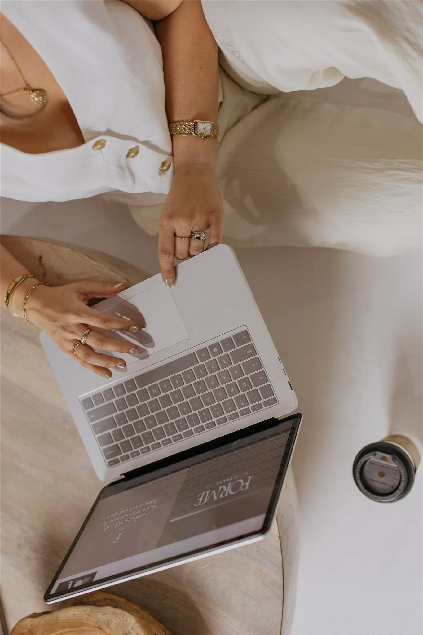 Person working on a laptop with a coffee cup nearby, wearing a white shirt with buttons and jewelry.