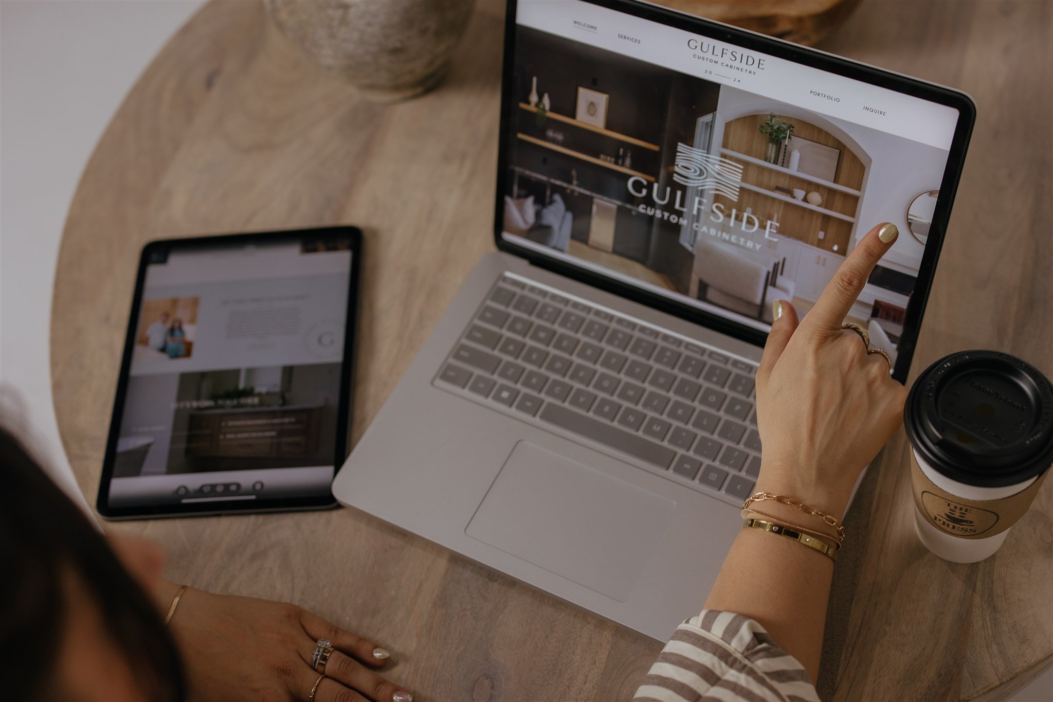 Person pointing at a laptop screen showing a website for Gulfside Custom Cabinetry, with a tablet and a coffee cup on a wooden table.