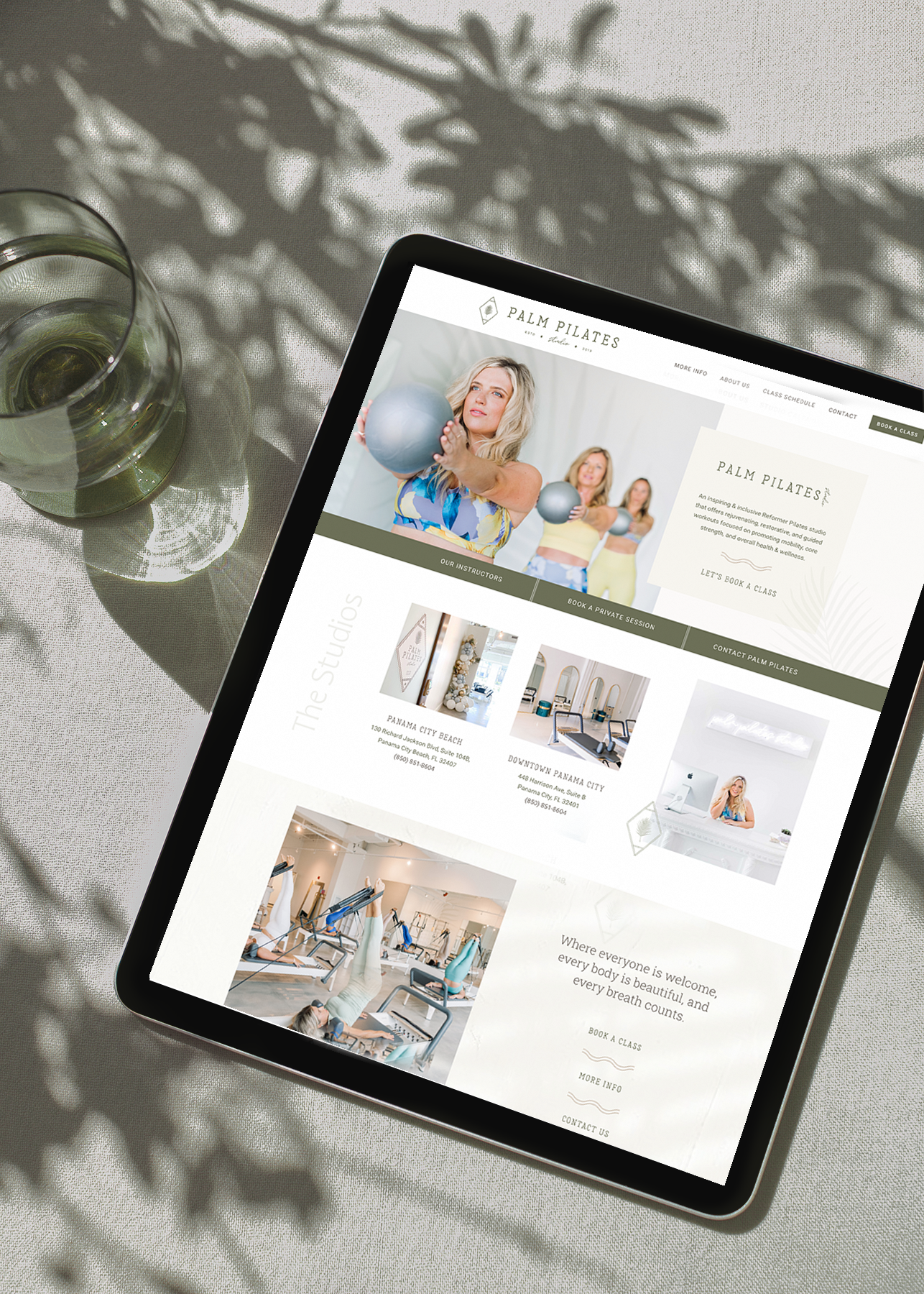 Tablet displaying a Pilates studio website with images of women exercising with exercise balls and equipment, placed on a table with a glass of water nearby.
