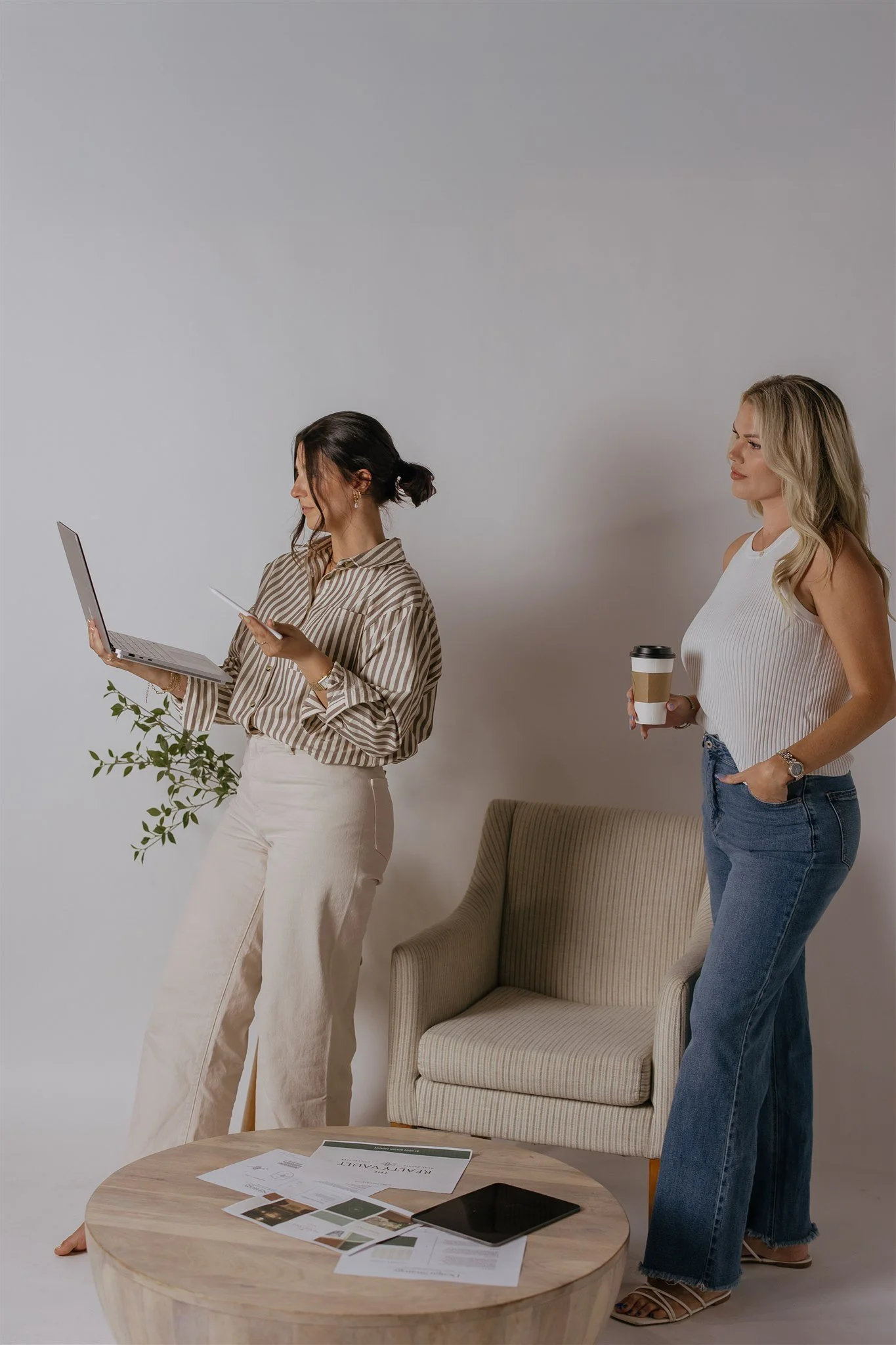 Two women in casual attire standing near a beige armchair, with a small round wooden table in front of them. One woman is holding a laptop and a coffee cup, and the other is holding a notebook and a branch of green leaves. There are papers and a tablet on the table.