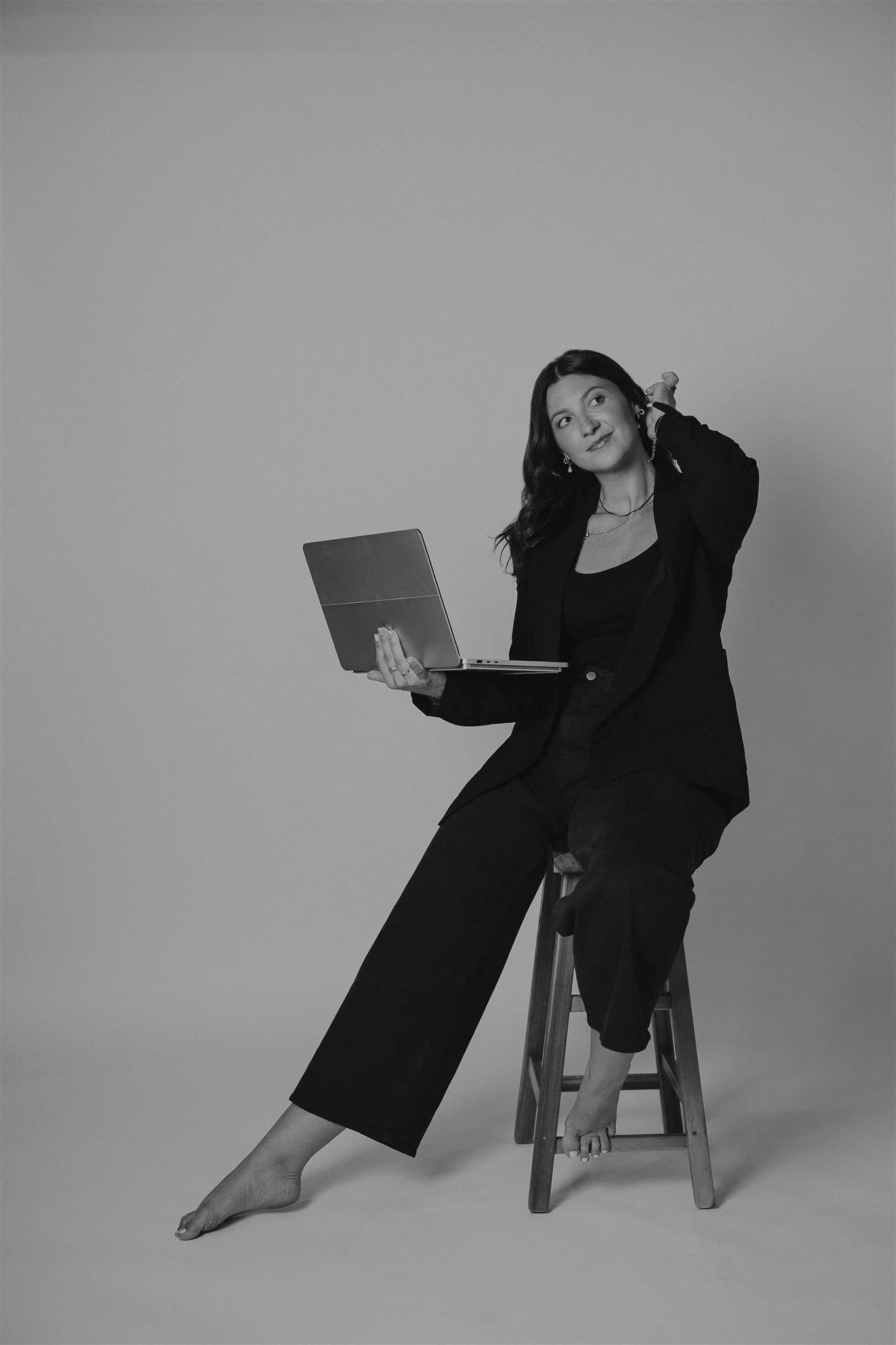 Katelyn Parsons, luxury brand & web designer, sitting on a wooden stool, holding a laptop with one hand, wearing a black blazer and pants, with one foot on the stool and the other resting on the ground, against a plain background.