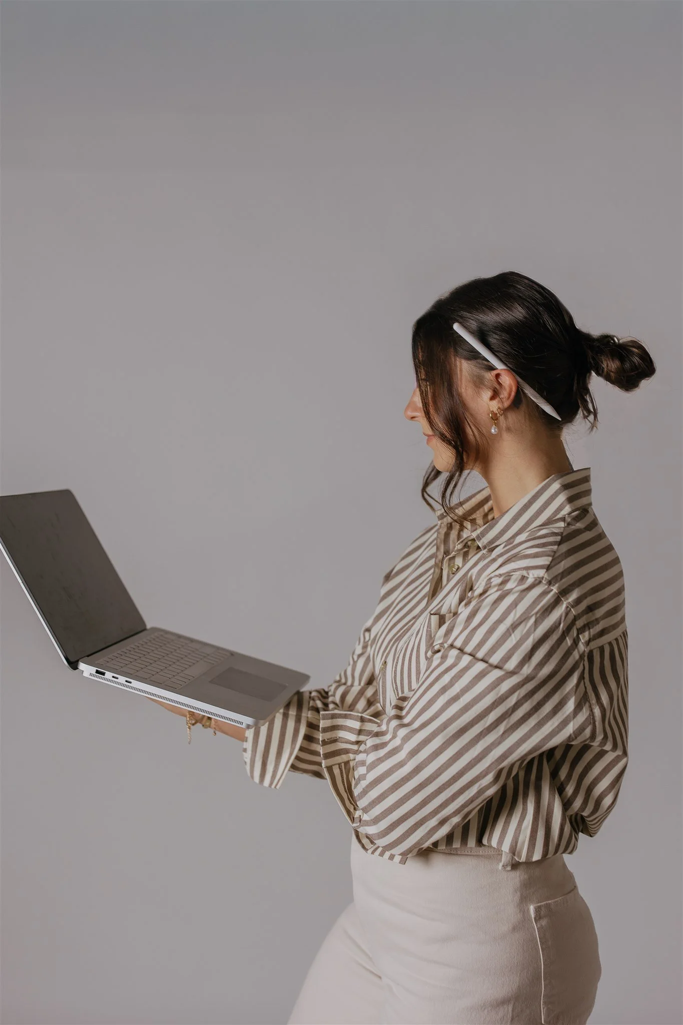 A woman with dark hair tied in a bun, wearing a beige and white striped shirt, holding a laptop and looking at the screen against a plain gray background.