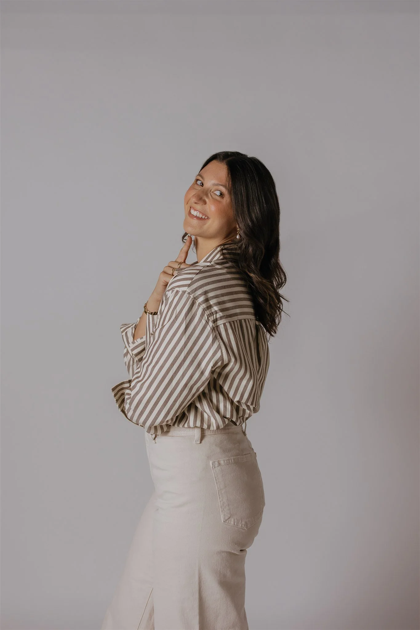 A woman with long wavy dark hair, wearing a striped beige and white shirt and beige pants, stands sideways with a smile and her right hand touching her chin against a light gray background.
