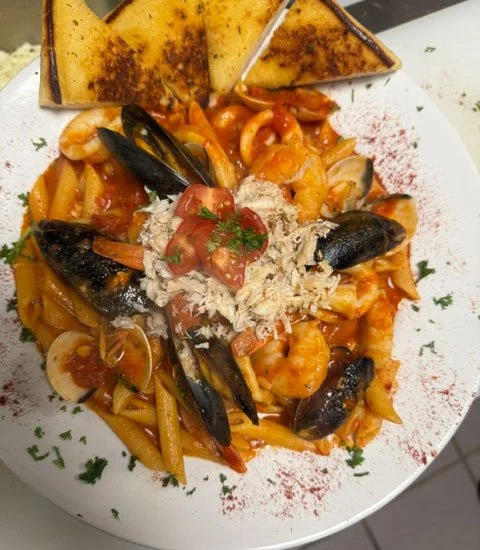 Seafood pasta dish with shrimp, mussels, and sliced calamari in tomato sauce, garnished with shredded cheese and fresh herbs, served with toasted bread.