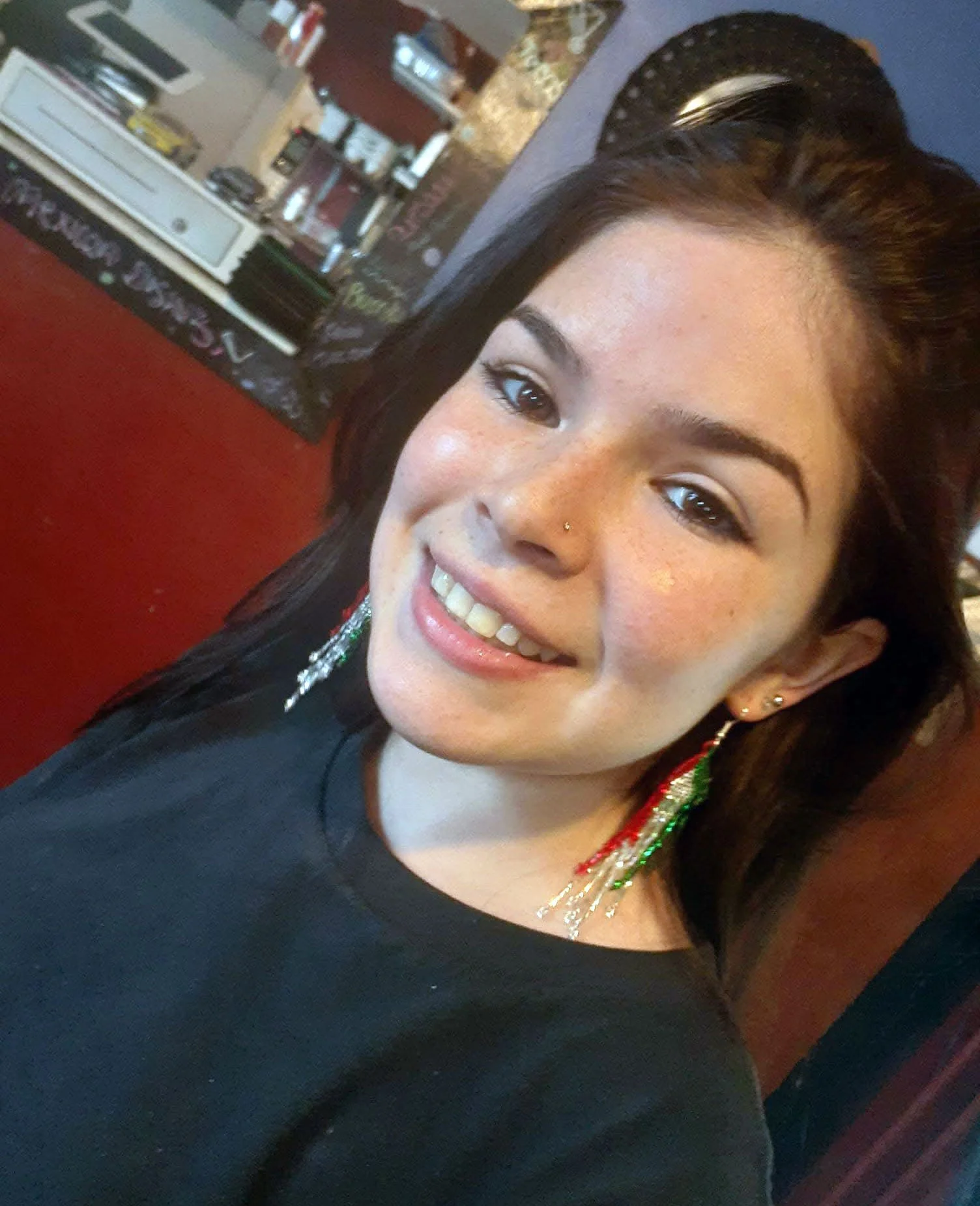 Close-up of a woman smiling with earrings, dark hair, and a nose piercing in a casual setting.
