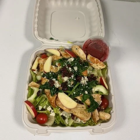 Salad with cherry tomatoes, apple slices, greens, cheese, croutons, and a side of red dressing in a takeout container.