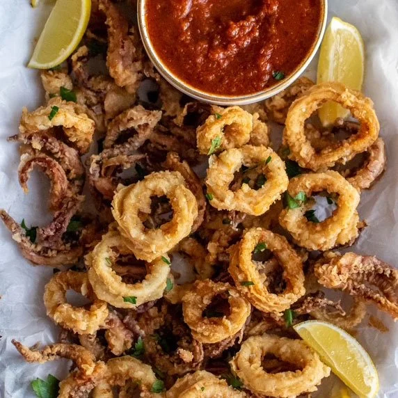 Fried calamari rings with lemon wedges and a small bowl of marinara sauce on a white paper-lined serving plate.