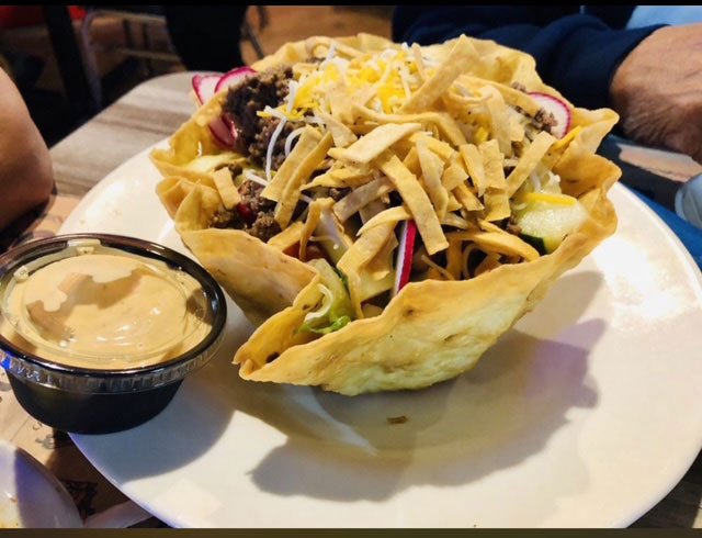 A taco salad in a crispy taco shell bowl topped with shredded cheese and vegetables, served with a side of creamy dressing.