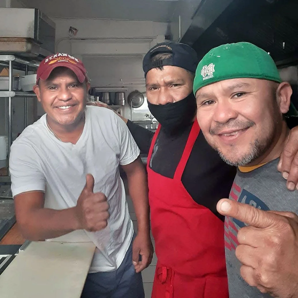 Three smiling men in a kitchen, one wearing a red cap and giving a thumbs-up, the second wearing a black cap and a face mask, the third wearing a green cap, all posing for a photo.