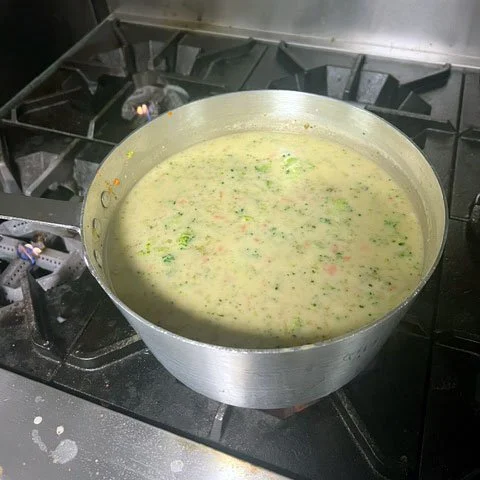 A large pot of creamy vegetable soup simmering on a gas stove with visible vegetables and herbs.