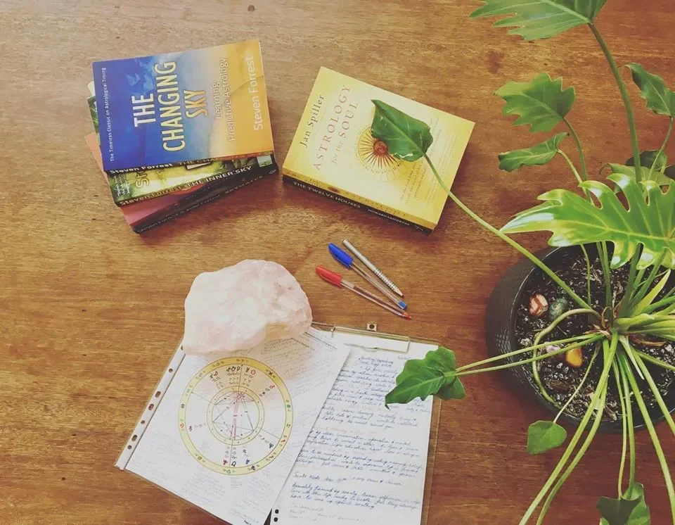 A wooden desk with several books, a potted plant, a pink Himalayan salt lamp, two pens, and handwritten notes with a zodiac wheel chart.