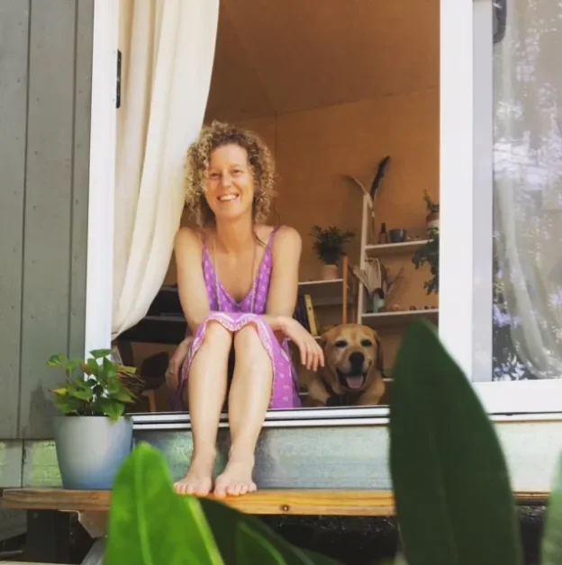 A woman with curly hair sitting on a windowsill next to a happy dog inside a house, with potted plants and shelves in the background.
