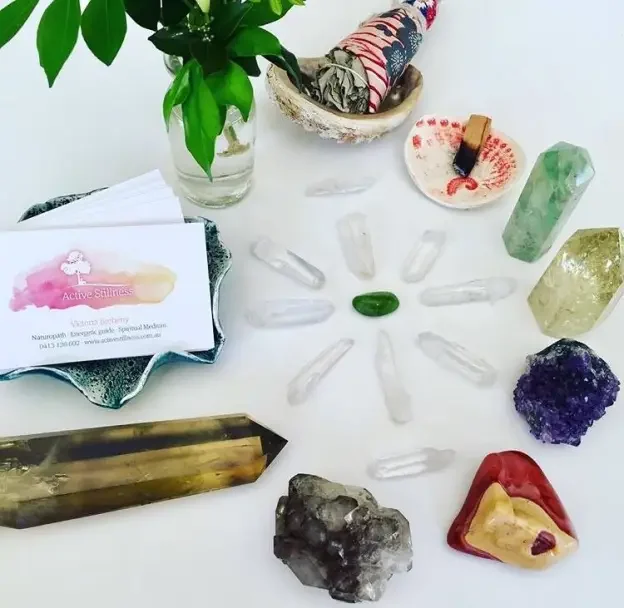 A table with various crystals, a green stone in the center, a small bowl with a colorful fabric item, a green plant in a glass vase, and a business card for Active Stillness.