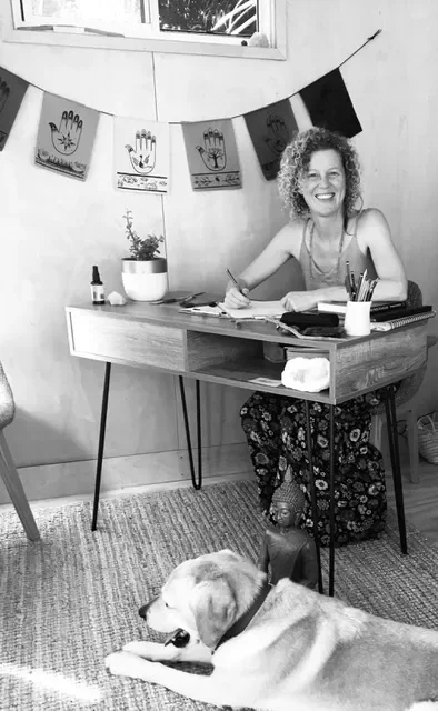 A woman with curly hair smiling and sitting at a desk with papers, pens, and a plant in a pot. Behind her, there is a string of artwork or cards hanging on a wall. A dog is lying on the floor in front of the desk.