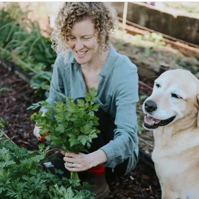 A woman with curly hair smiling as she harvests herbs in a garden, with a happy Labrador Retriever beside her.