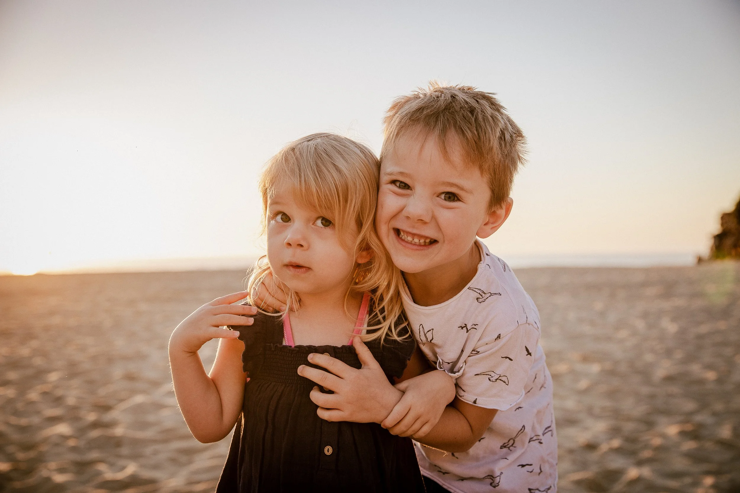 A happy young boy with short, tousled hair is hugging a young girl with long, light-colored hair. They are outdoors on a beach during sunset, with the ocean and a slightly blurred landmass or cliff in the background.