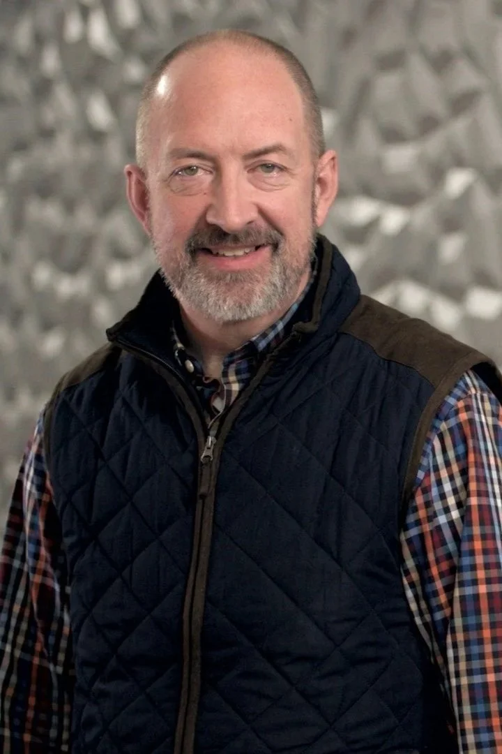 A middle-aged man with a beard and short hair, smiling, wearing a plaid shirt and a dark quilted vest, against a blurred gray and white background.