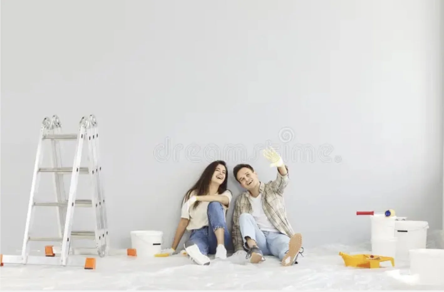 Two people, a woman and a man, sitting on the floor with painting supplies, smiling, with a ladder and painting tools around them, in a room they are painting.