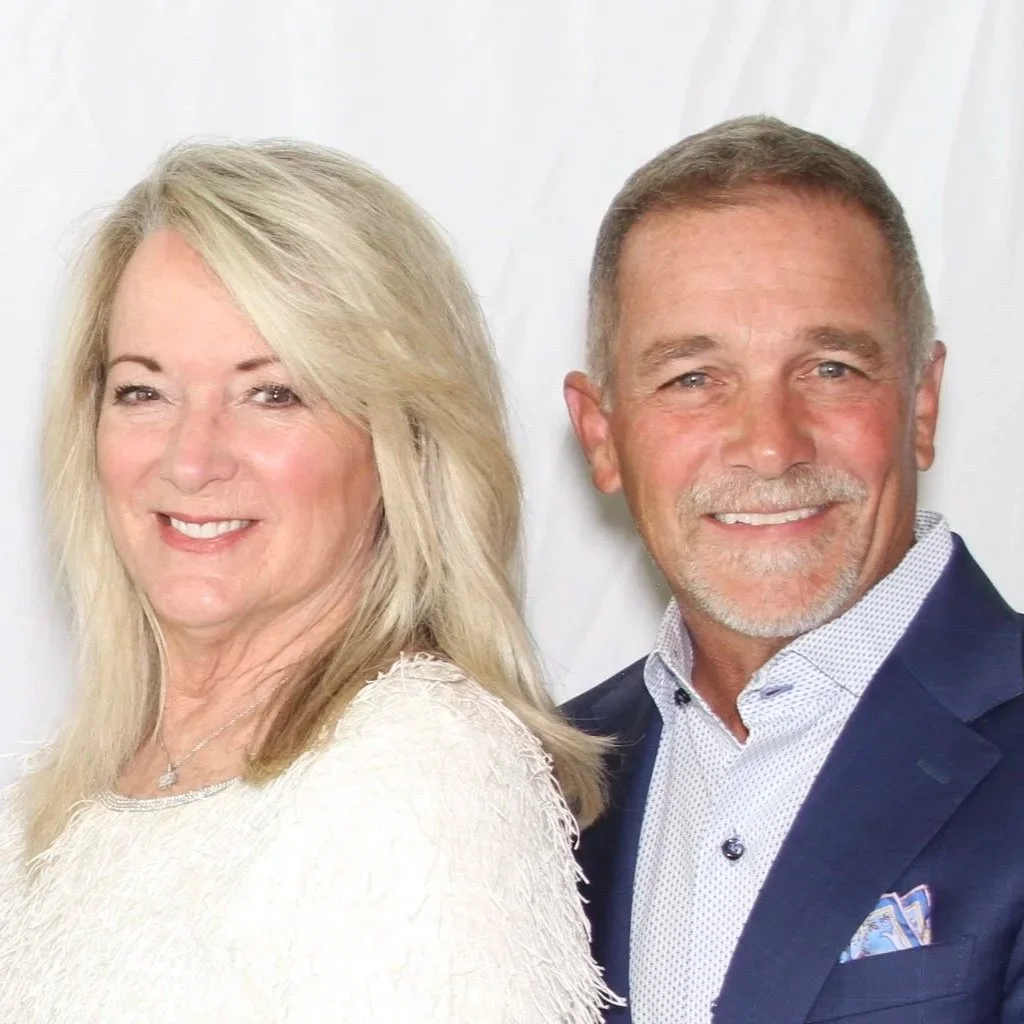 A smiling middle-aged couple, woman with long blonde hair wearing a white dress, man with short gray hair and beard wearing a navy suit and white shirt, standing together against a plain white backdrop.