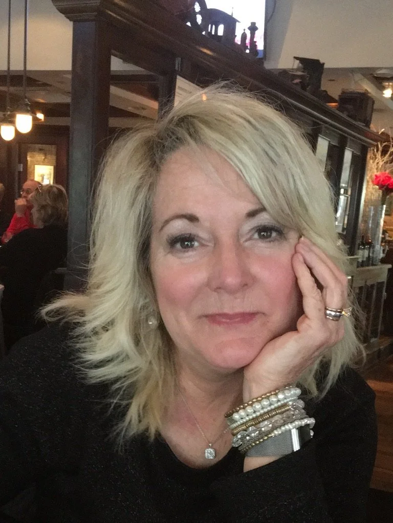 A woman with blonde hair and light skin, sitting indoors, resting her face on her hand, smiling softly, wearing jewelry, and in a cozy setting with warm lighting.