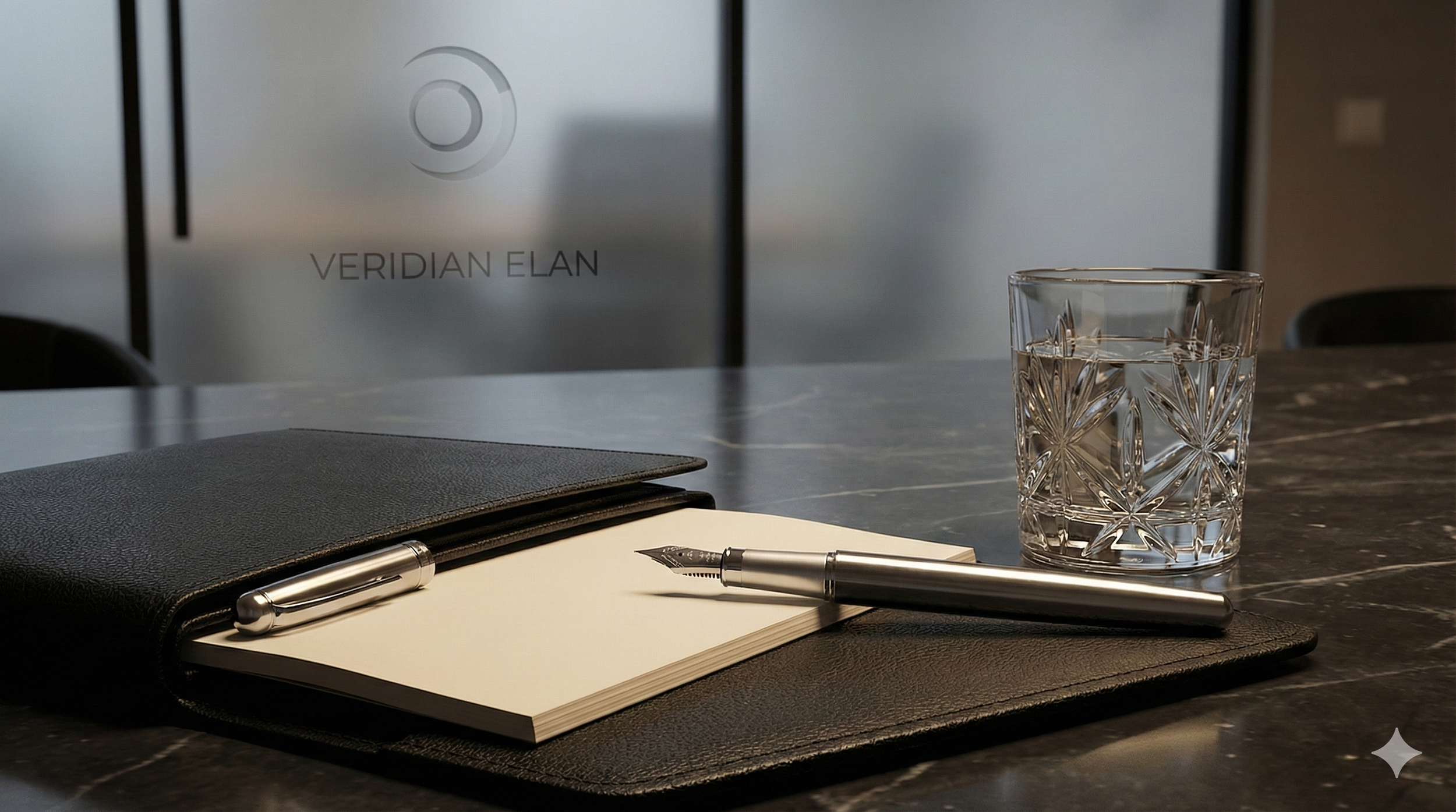 A black notebook, silver fountain pen, empty notepad, and patterned glass of water on a dark marble table in a modern office setting.