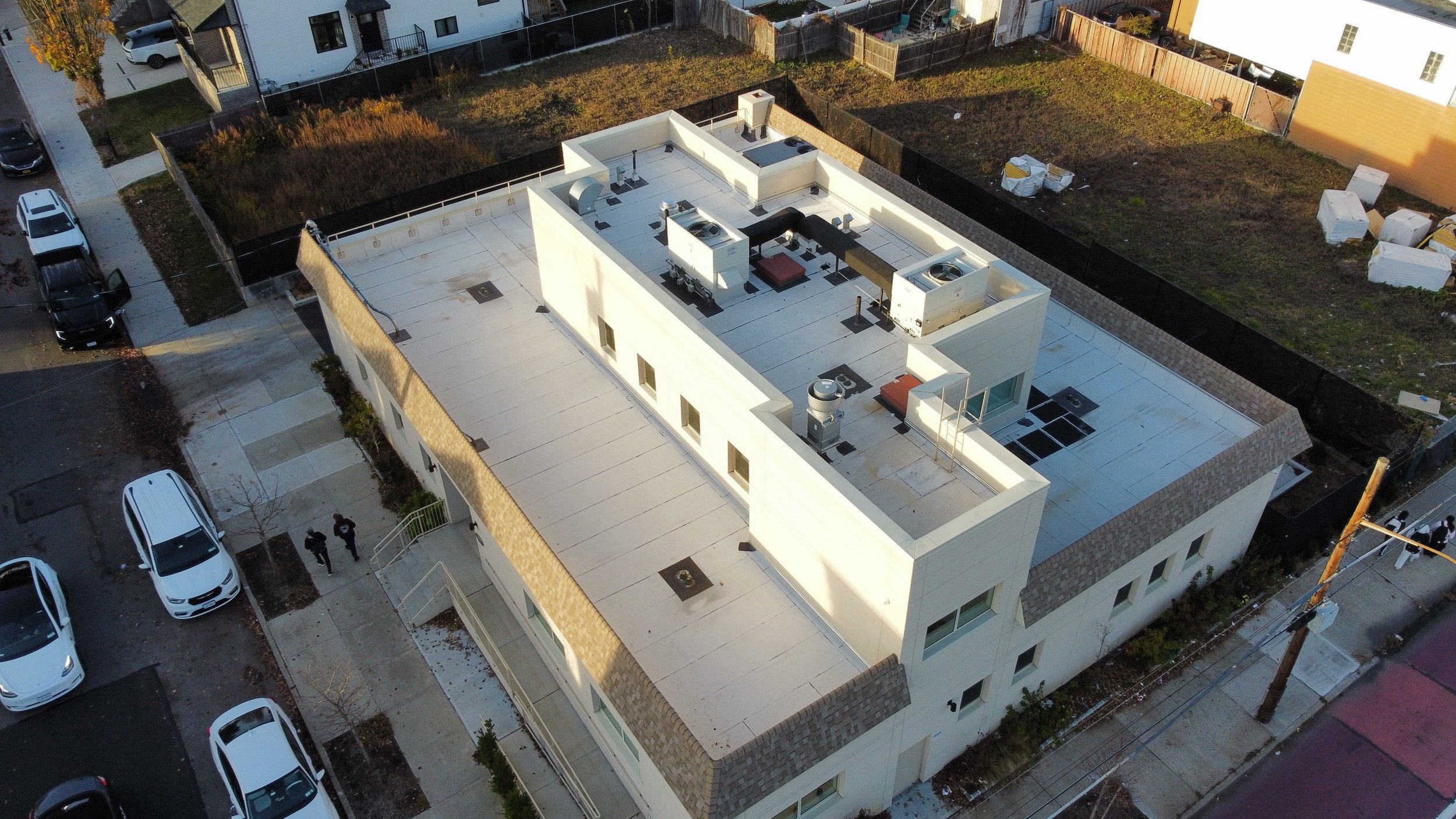Aerial view of a modern white multi-story building with a flat roof, surrounded by parked cars and a neighborhood with houses and empty lots.