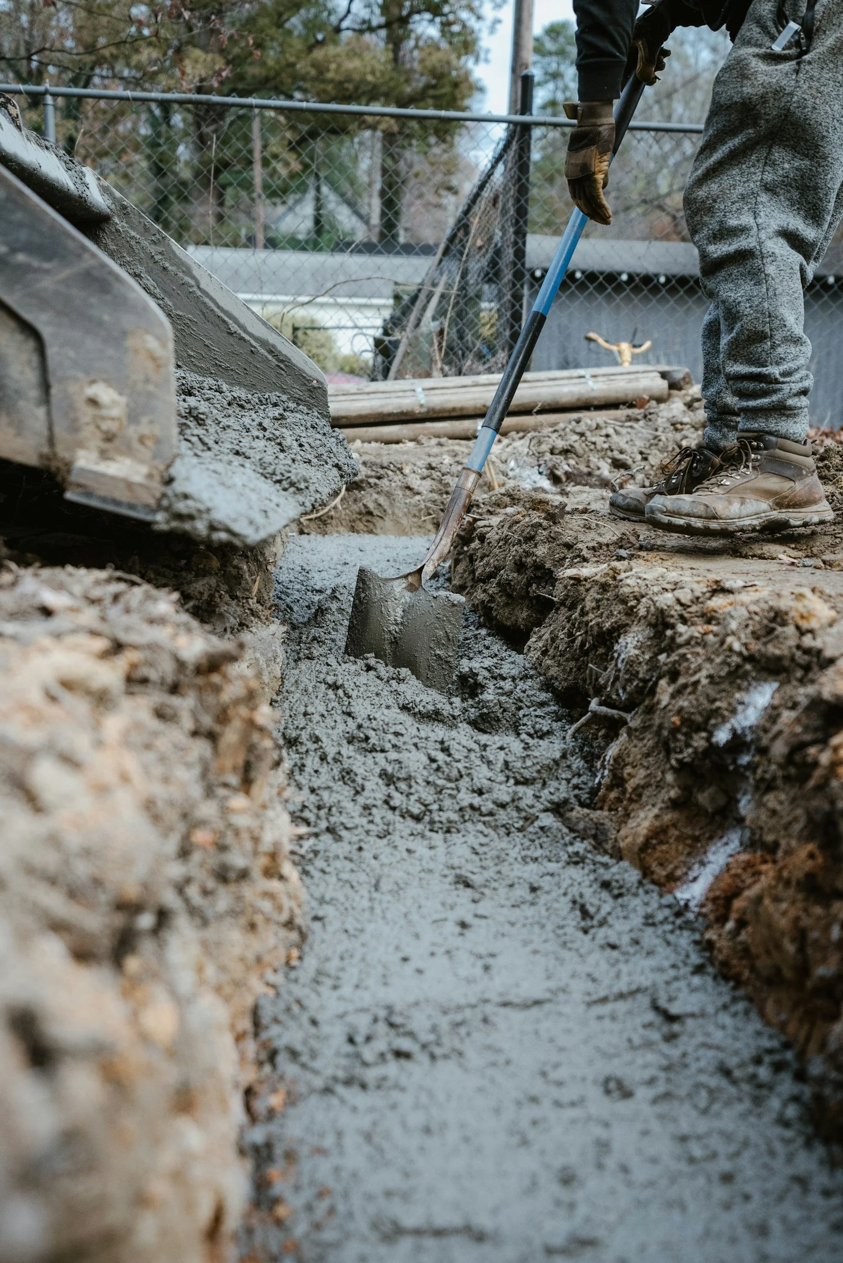 Person wearing work boots and gloves using a shovel to spread concrete in a trench at a construction site, with a chain-link fence and trees in the background.
