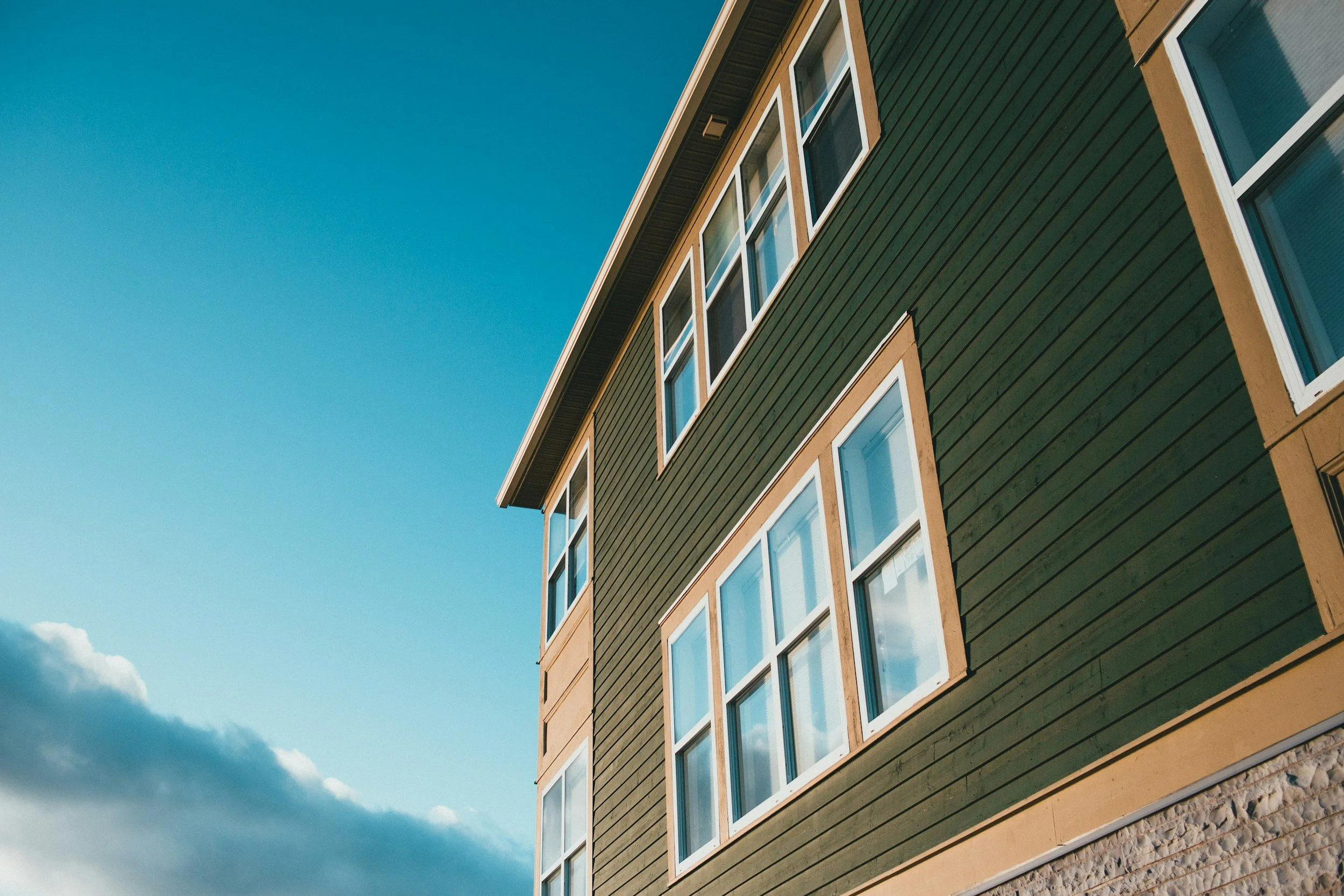 Low-angle view of a modern green wooden apartment building with multiple white-framed windows against a blue sky with some clouds.