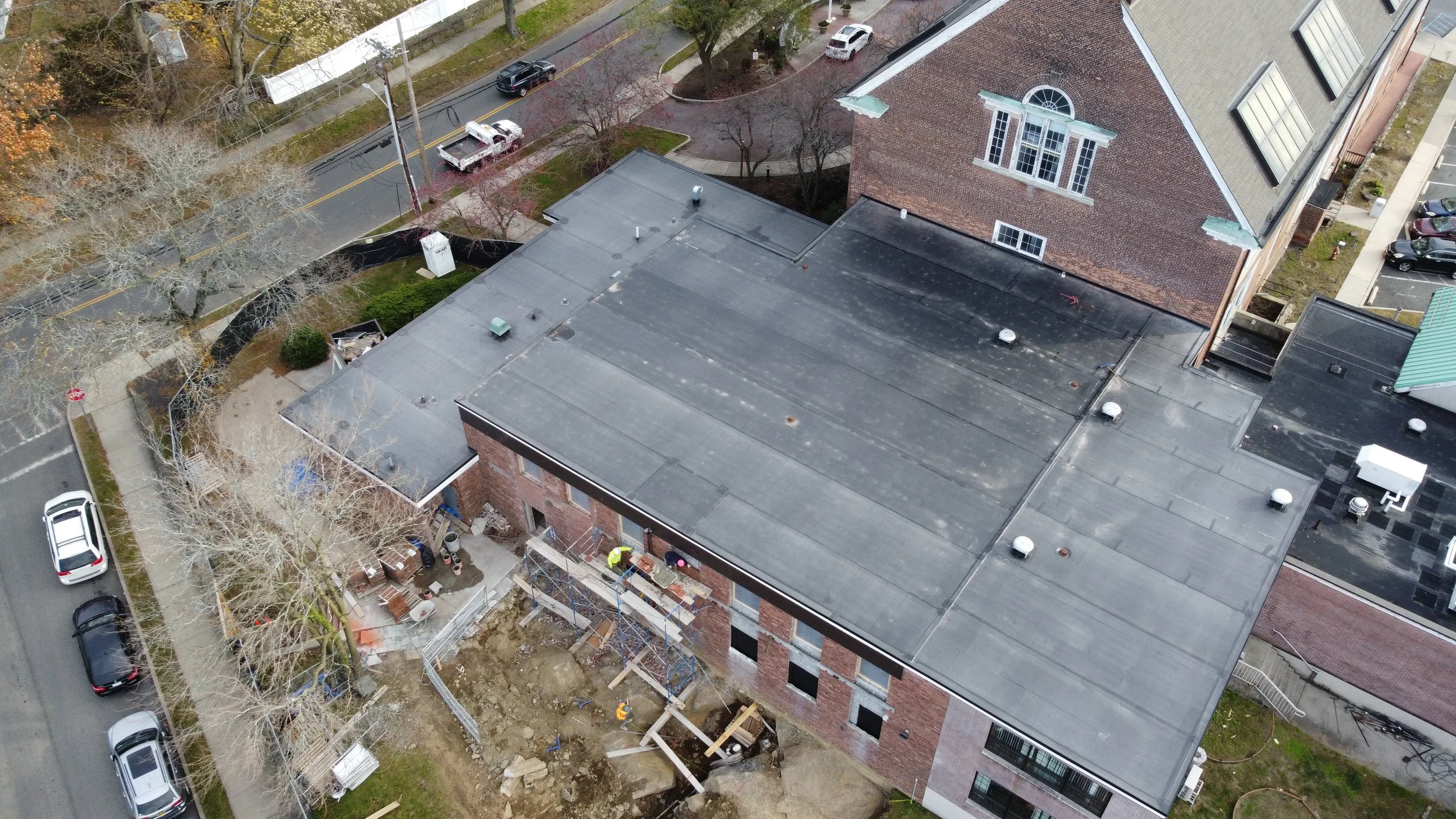 An aerial view of a construction site on a building with workers, scaffolding, and an exposed ground area, surrounded by parked cars and nearby residential buildings.