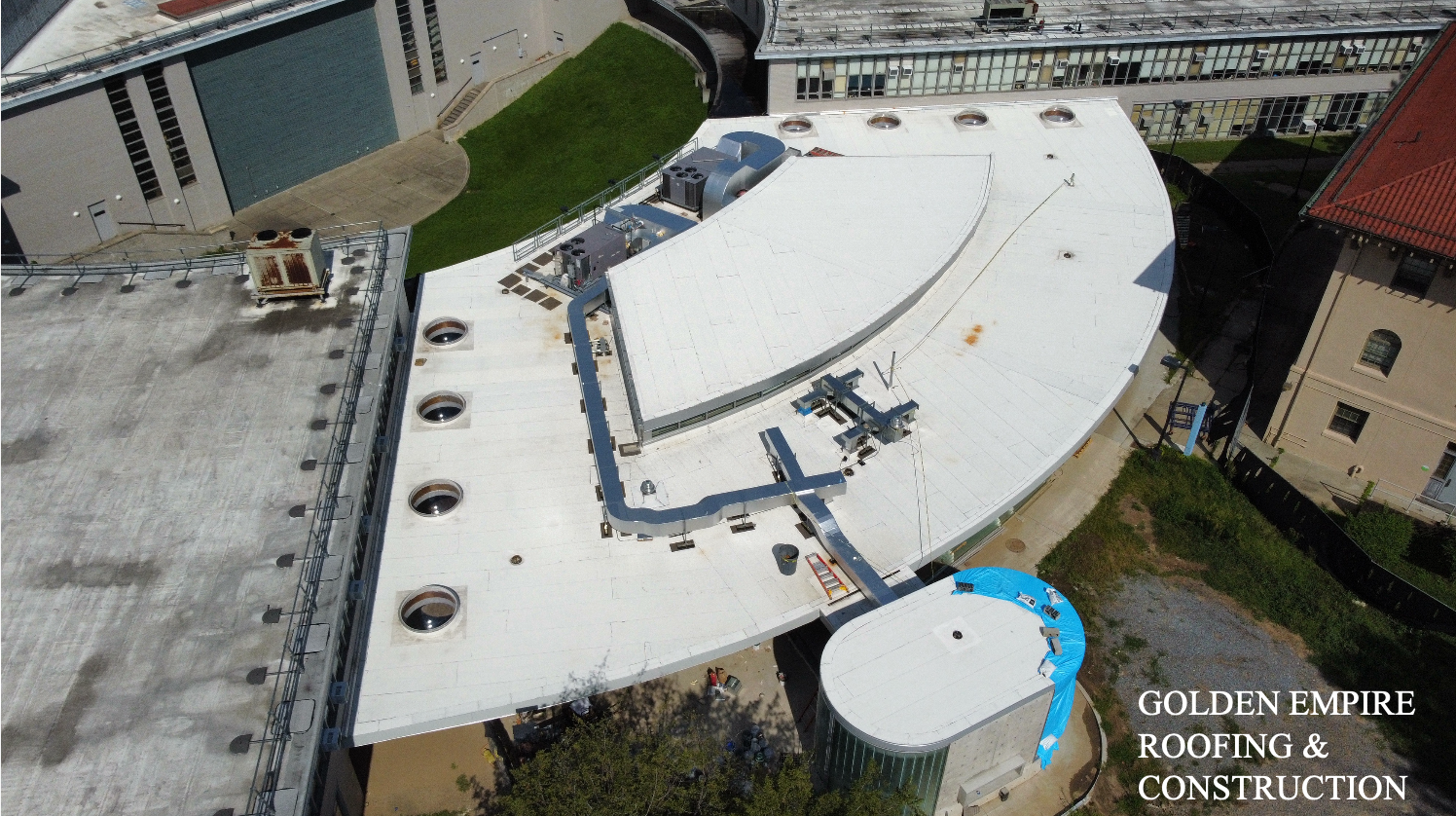 An aerial view of a building's roof showing ventilation units, a curved structure, various piping and equipment, and surrounding buildings with some green patches.