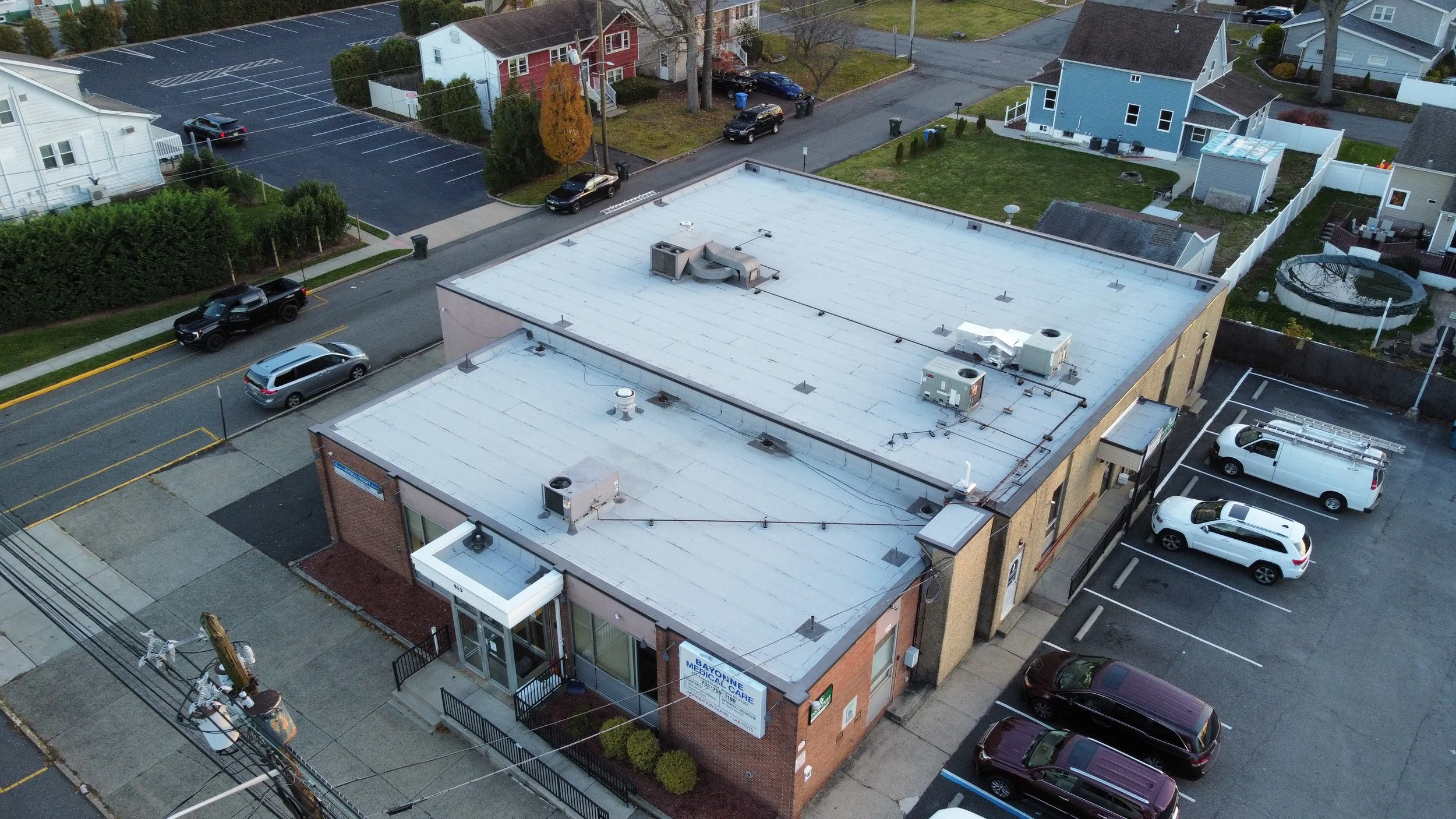 Aerial view of a brick commercial building with a flat roof, parking lot with several cars, and neighboring residential houses with fences, trees, and yards.