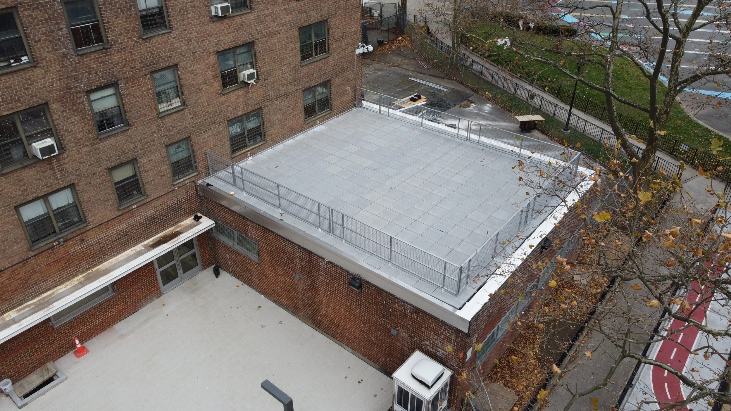 An aerial view of a city rooftop with a fenced basketball or tennis court, adjacent brick buildings, leafless trees, a parked car, a sidewalk, and a nearby parking lot.