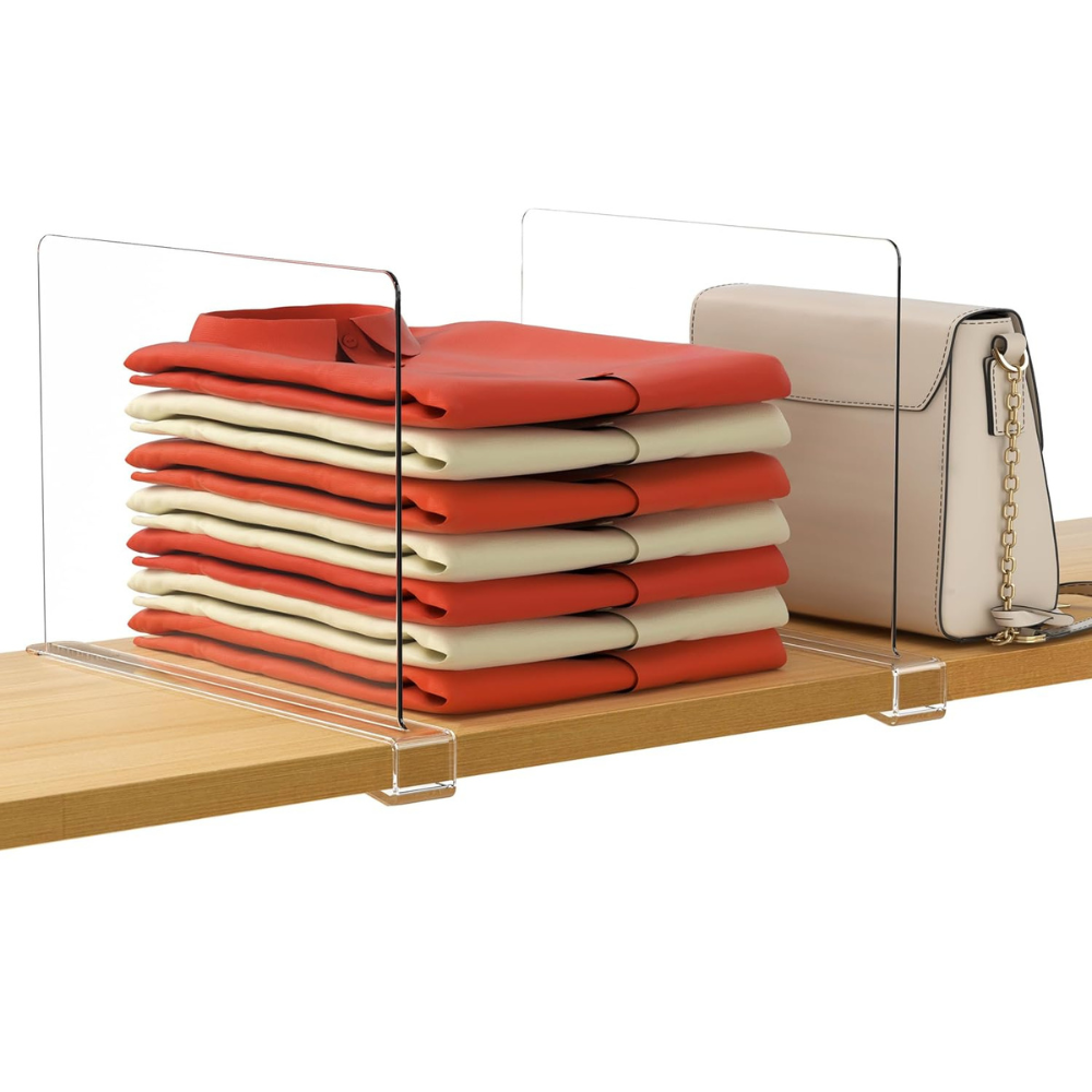 Stack of folded clothes, a beige handbag with a chain strap, separated by a transparent acrylic divider on a wooden shelf