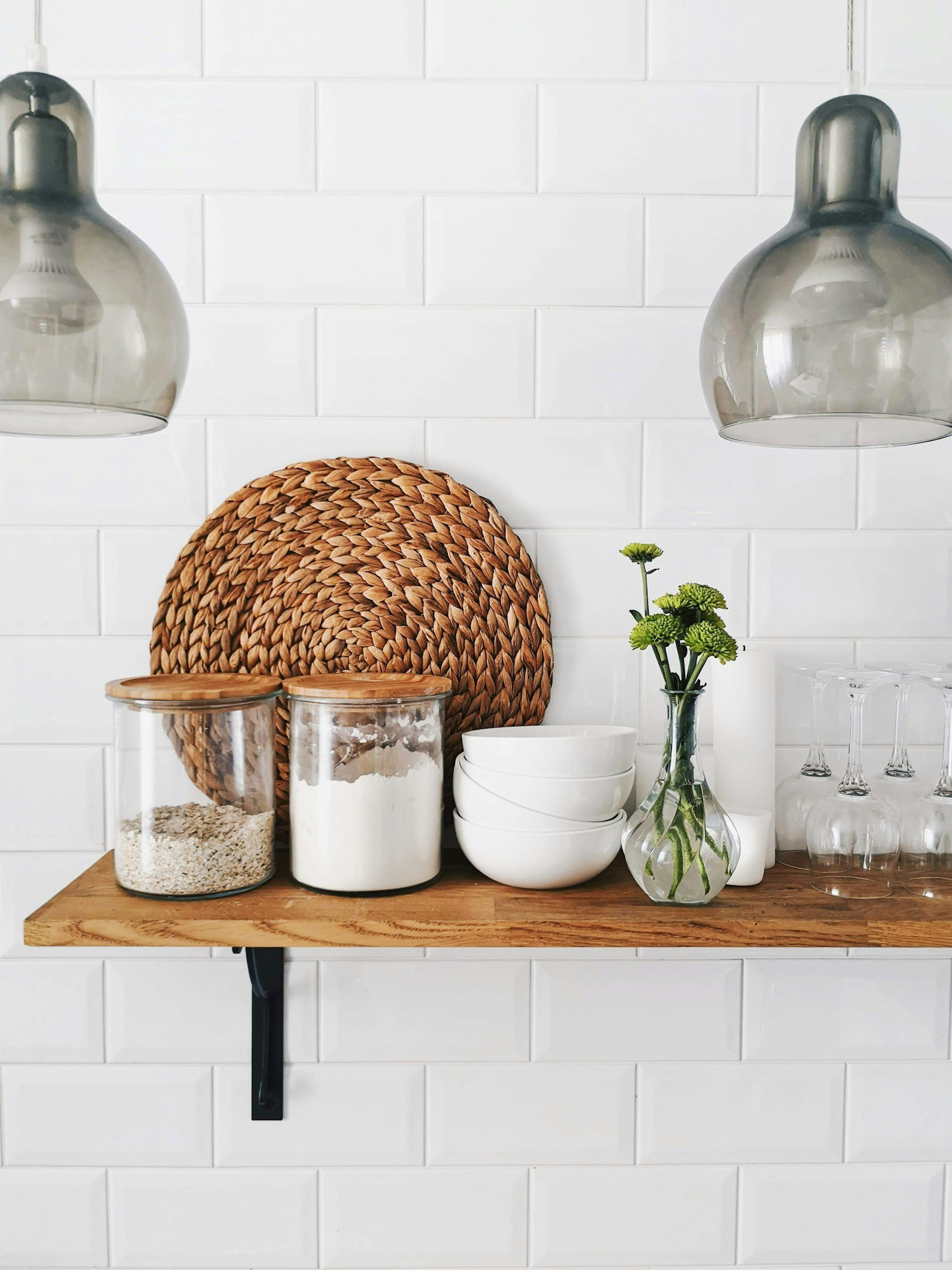A wooden wall shelf mounted on a white tiled wall, holding glass jars, white bowls, a glass vase with green flowers, and clear glassware, with decorative woven placemat and black metal brackets.