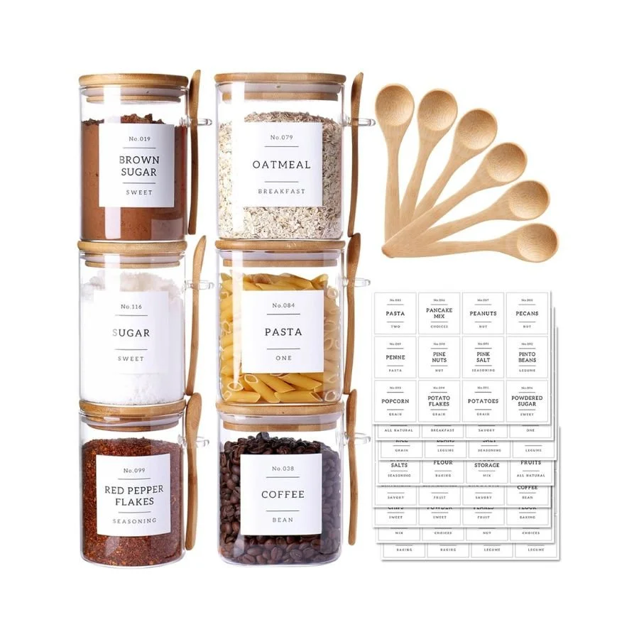 Six glass jars filled with baking ingredients labeled as brown sugar, oatmeal, sugar, pasta, red pepper flakes, and coffee, arranged in two columns, with six wooden spoons and baking labels on paper nearby.