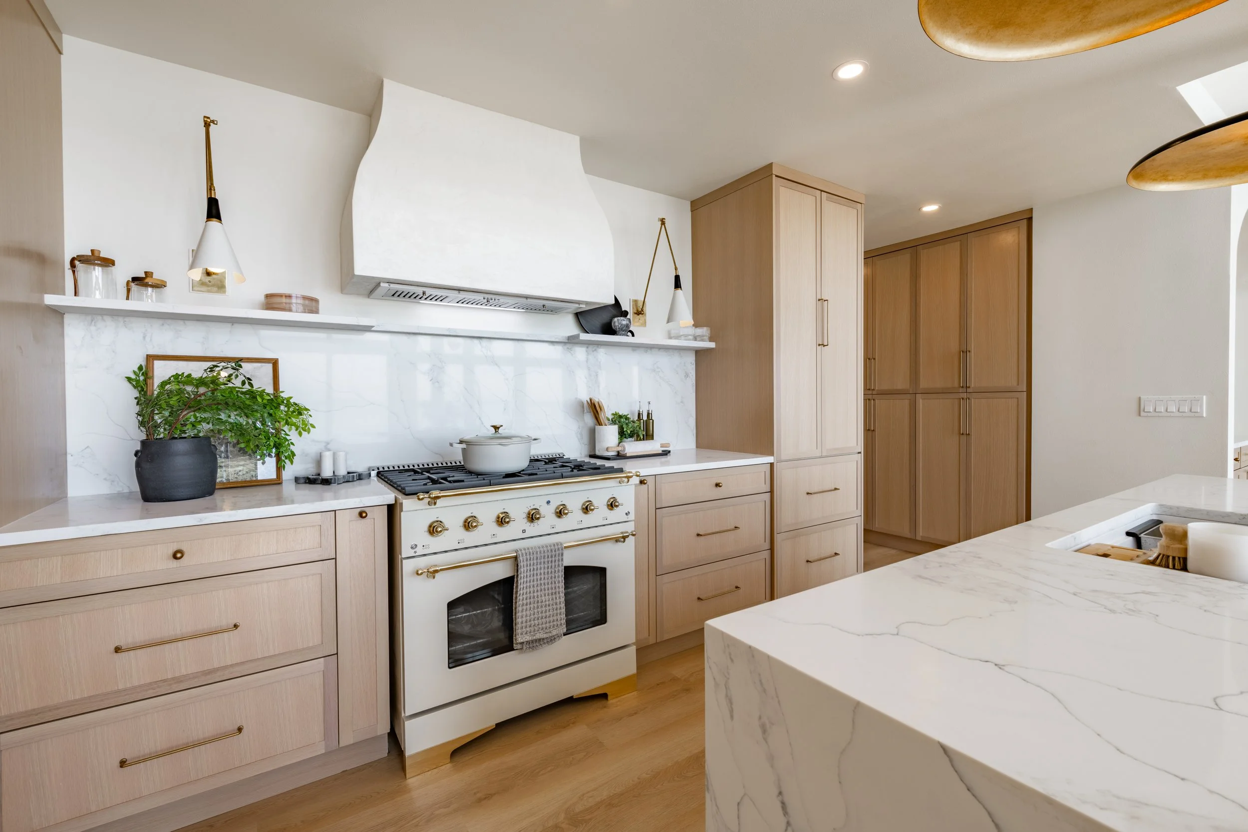 Modern kitchen with light wood cabinets, white marble countertops, a white oven, and minimal decor including a green plant, glass jars, and gold accents.