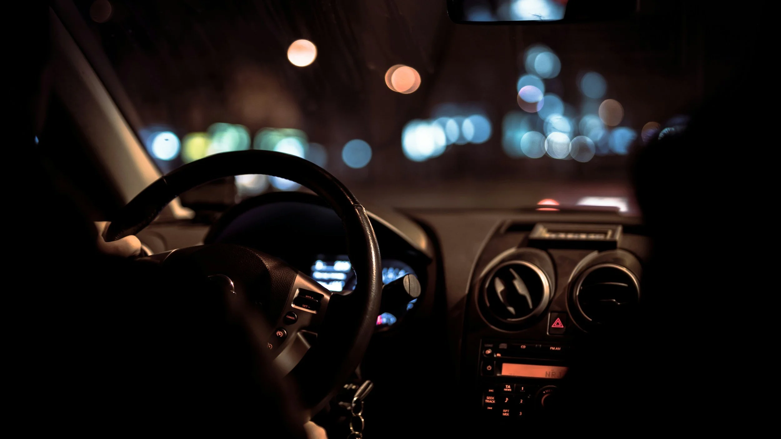 View of a car interior at night, showing the steering wheel, dashboard with illuminated controls, and blurry city lights outside through the windshield.