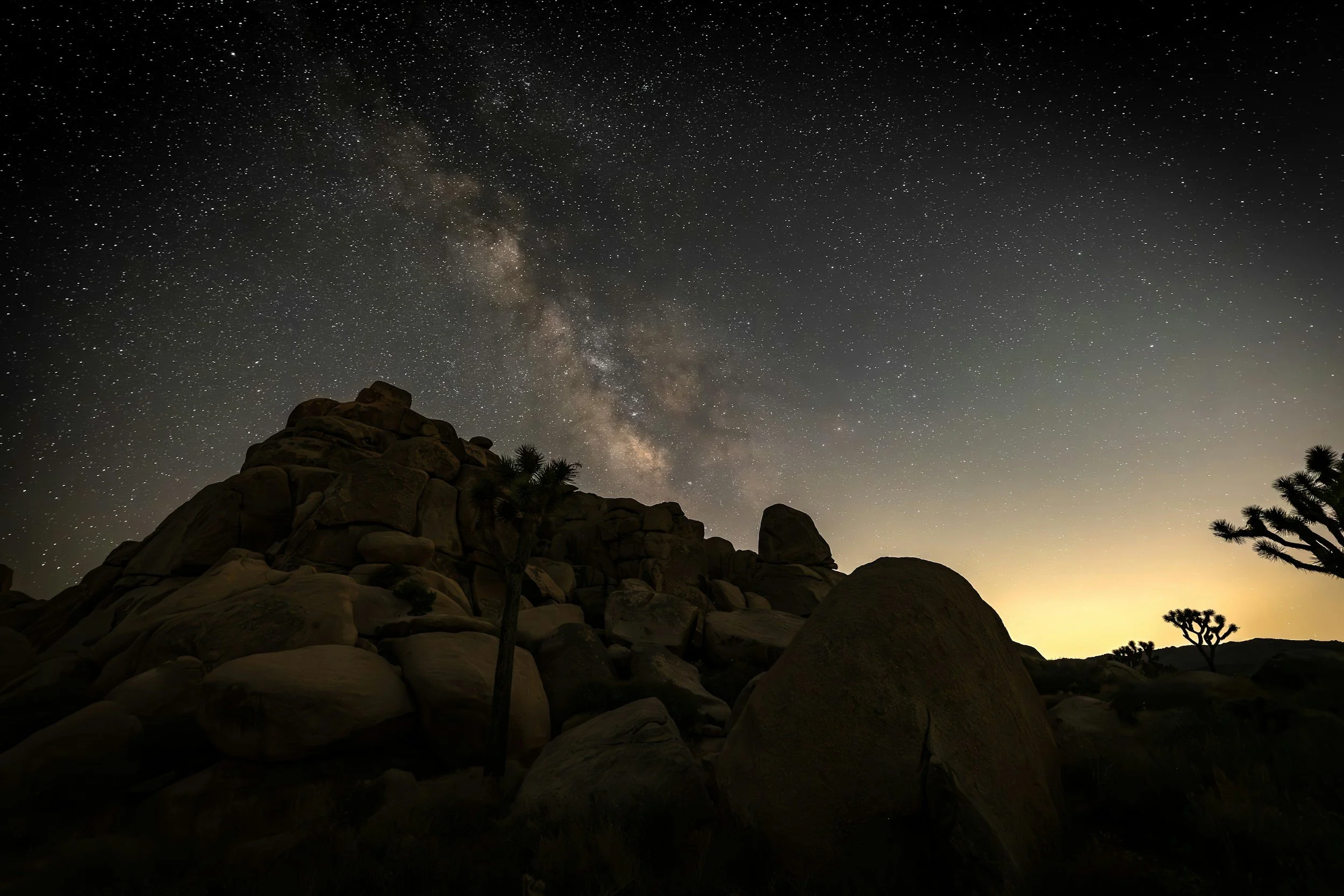 Night sky filled with stars and the Milky Way galaxy over a rocky desert landscape with Joshua trees.