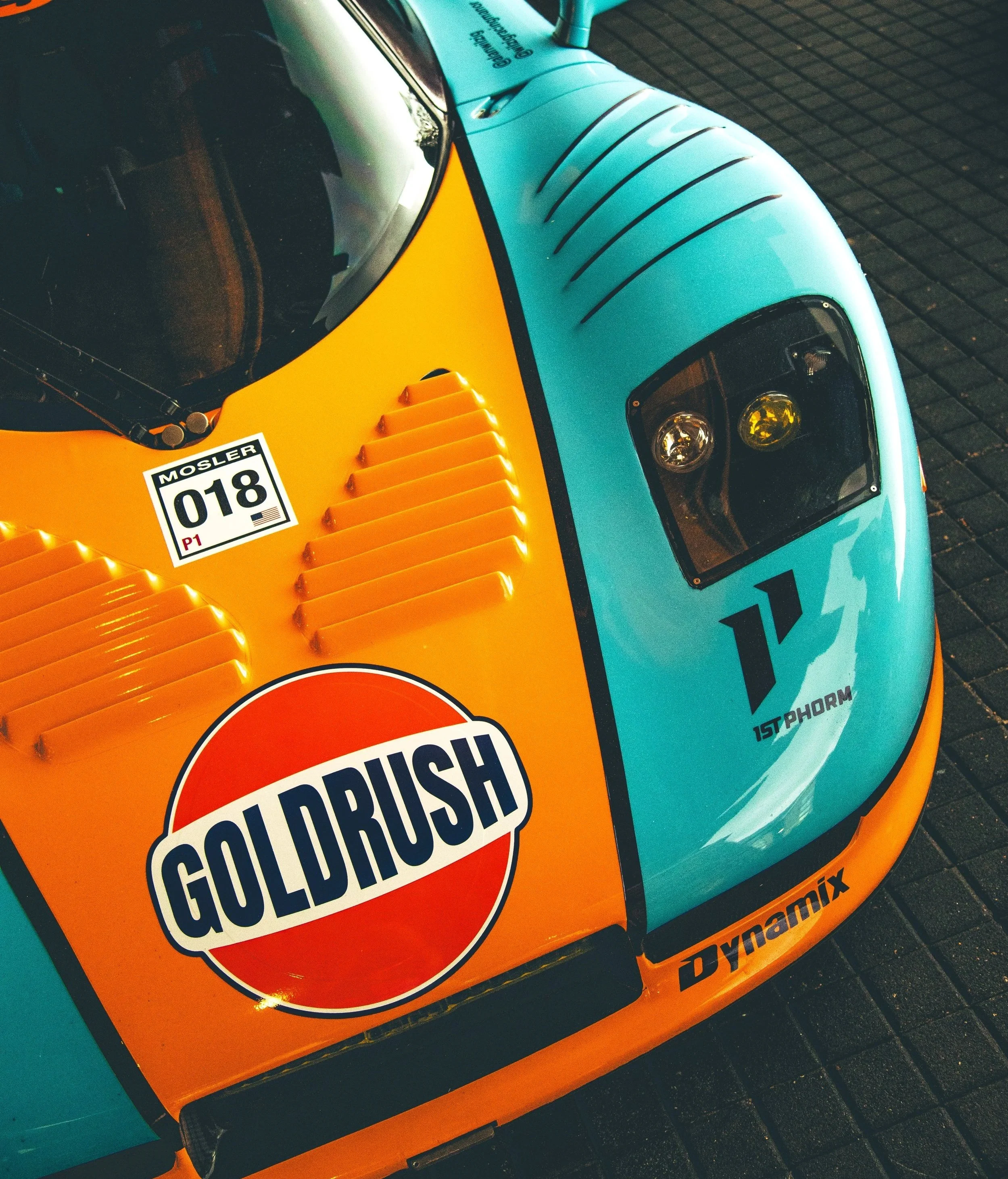 Close-up of a vintage racing car with a bright orange and light blue color scheme. The car features prominent logos, including a 'Gold Rush' logo on the orange section and a '1st Phorm' logo on the blue section. The car has a racing number '018' sticker with the name 'Mosler' and a small American flag. The front of the car shows vents and headlights.